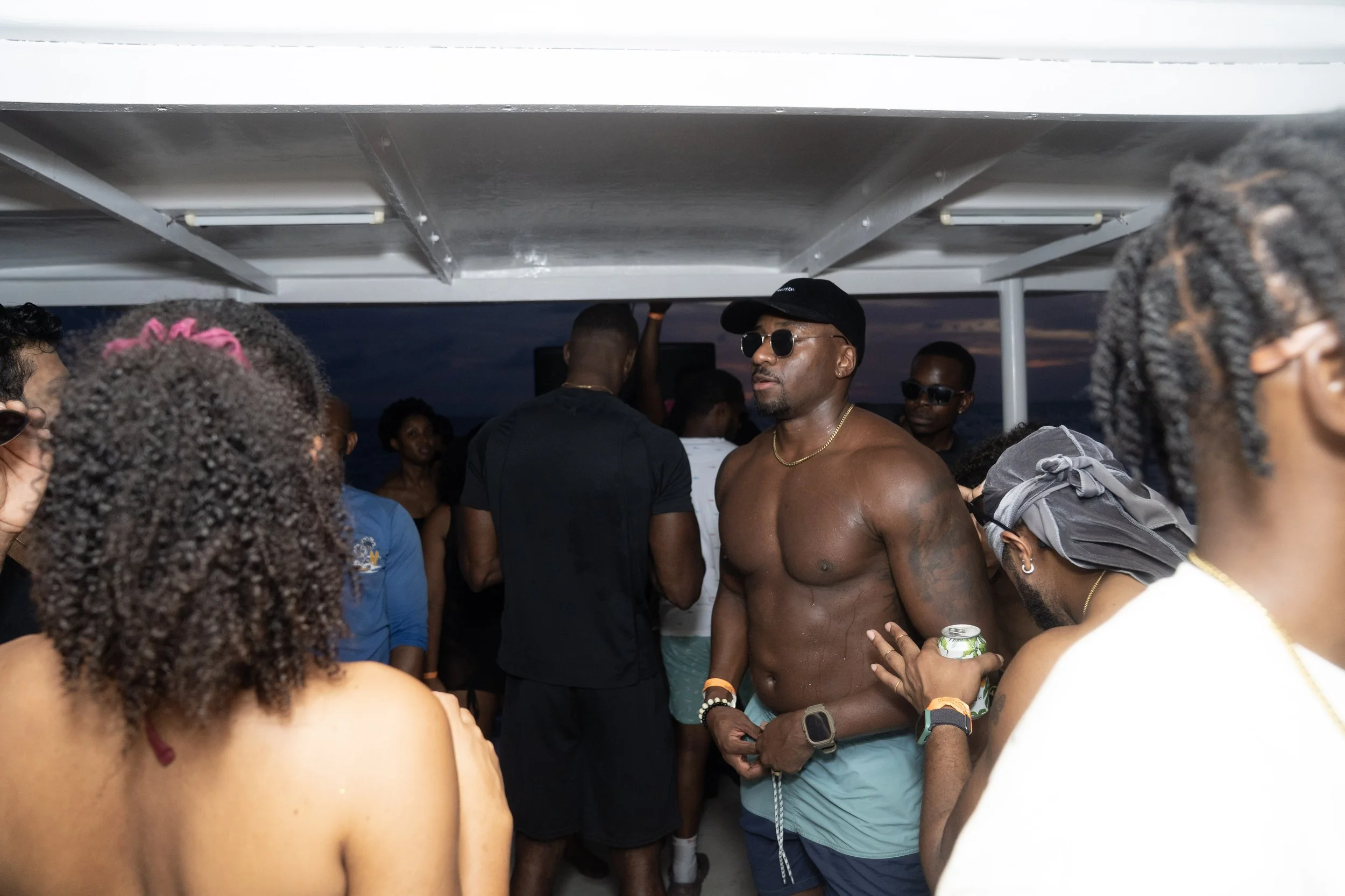Group of people enjoying a boat party at sunset, some wearing sunglasses, casual clothing, and swimwear, with a shirtless man in the center holding a drink.