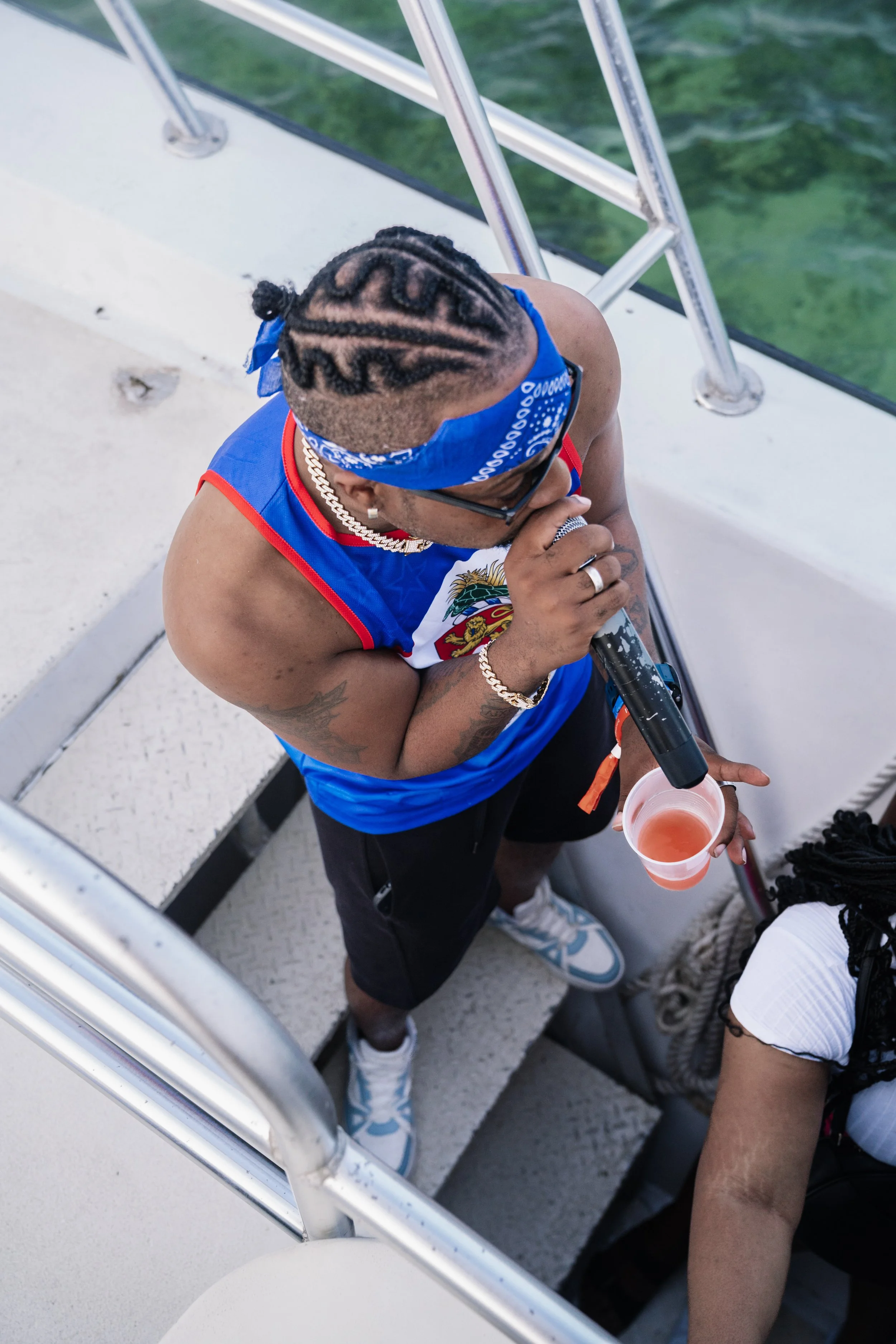 Man with braided hair, sunglasses, and jewelry singing into a microphone while holding a cup, on a boat with water in the background.