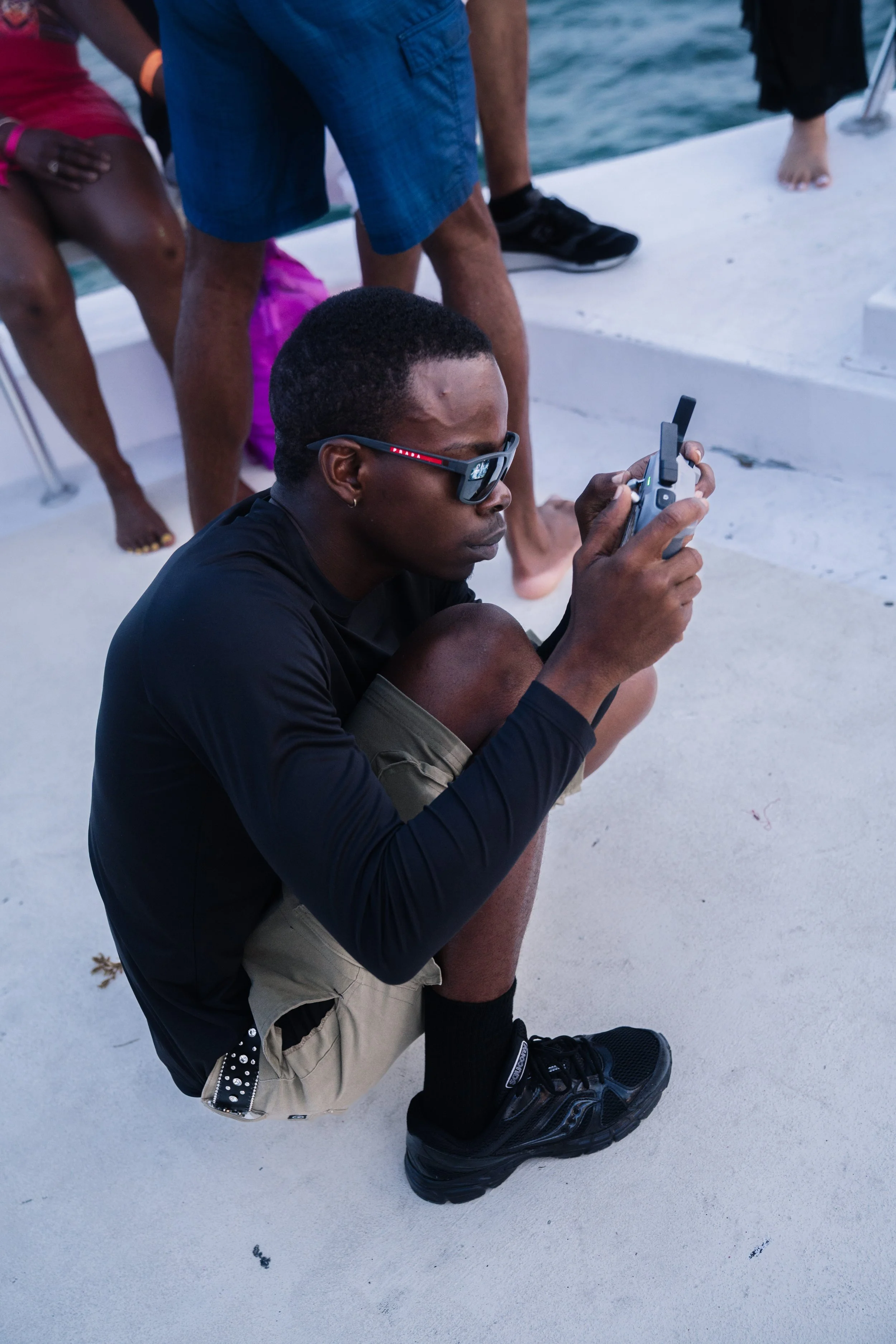 A man wearing sunglasses crouches on a boat deck, looking at a handheld device, with other people standing and sitting nearby on the boat.