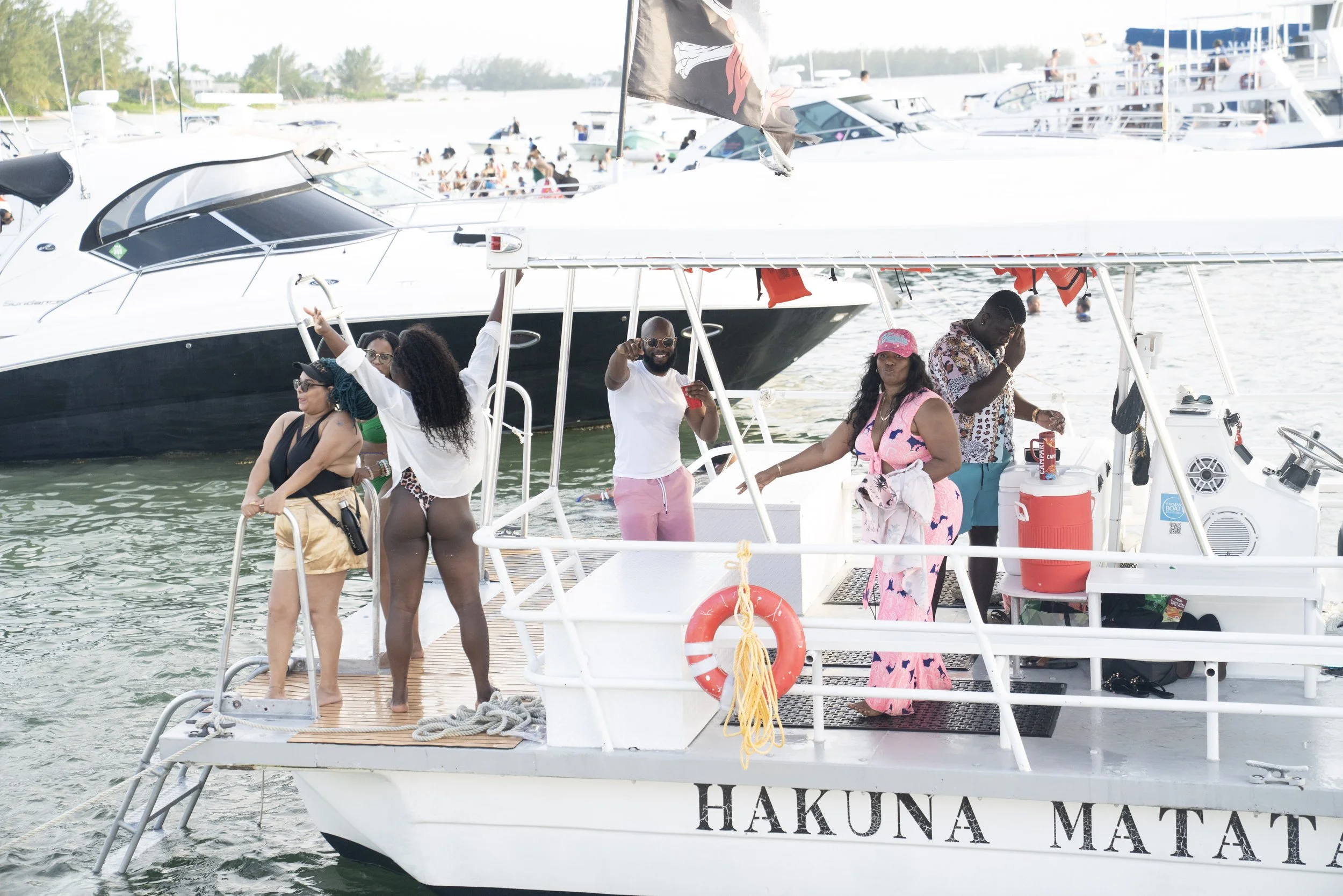 People enjoying a boat party on a yacht with “Hakuna Matata” written on the side, in a marina with other boats in the background.