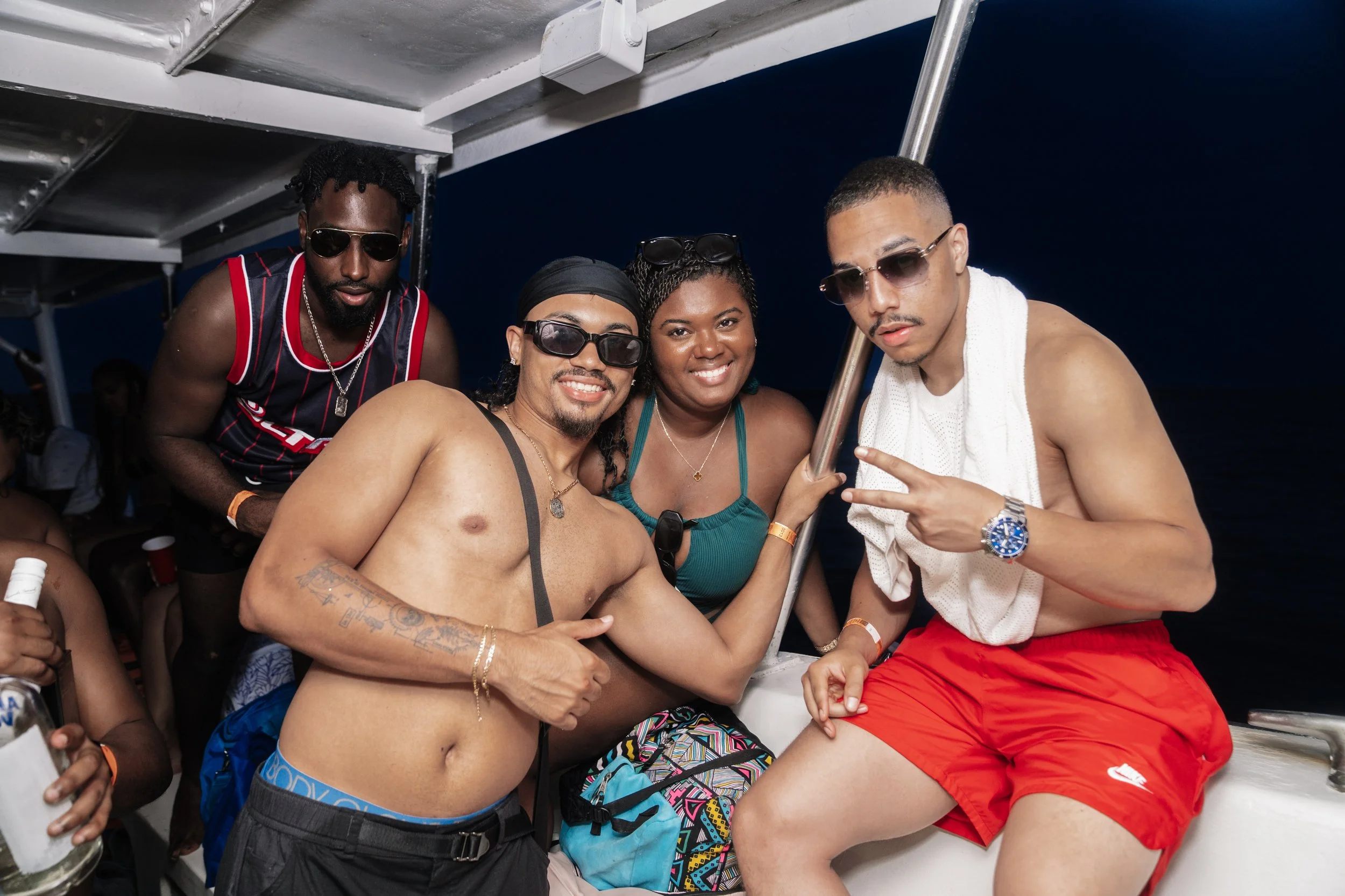 Group of friends on a boat enjoying a night out, some wearing sunglasses, in casual summer attire, with a dark water background.