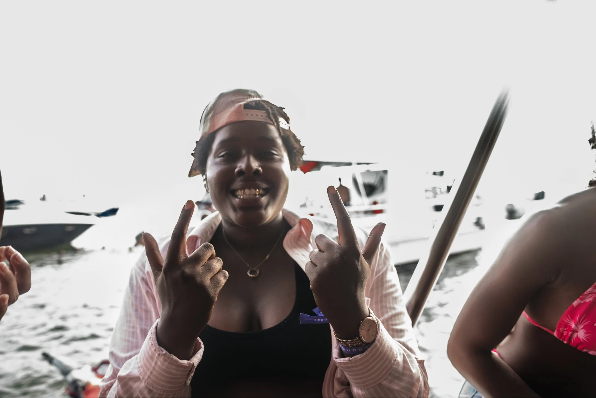 A young person smiling at the camera making the 'rock on' hand gesture, near boats on water.