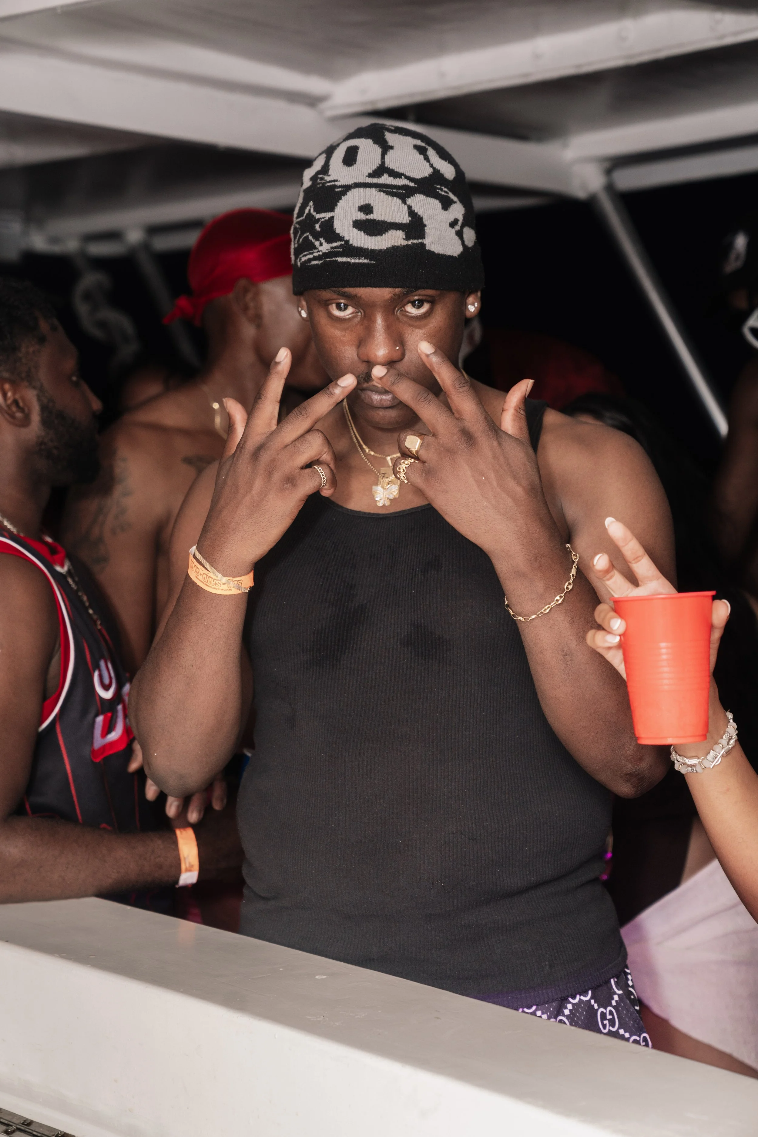 A person with a black beanie, wearing a black tank top, standing among a group of people at a party holding a red cup, with other partygoers in the background.