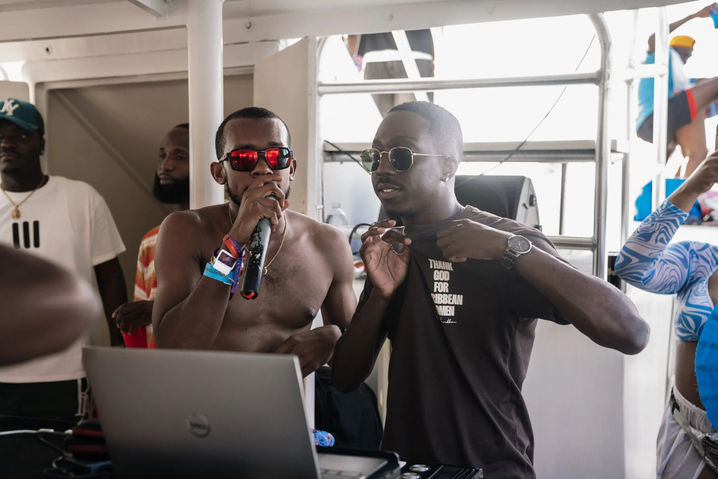 NXTGEN SOUND wearing sunglasses at a party, one shirtless with a microphone, the other wearing a black T-shirt with white text, standing behind a DJ setup, with multiple people in the background.