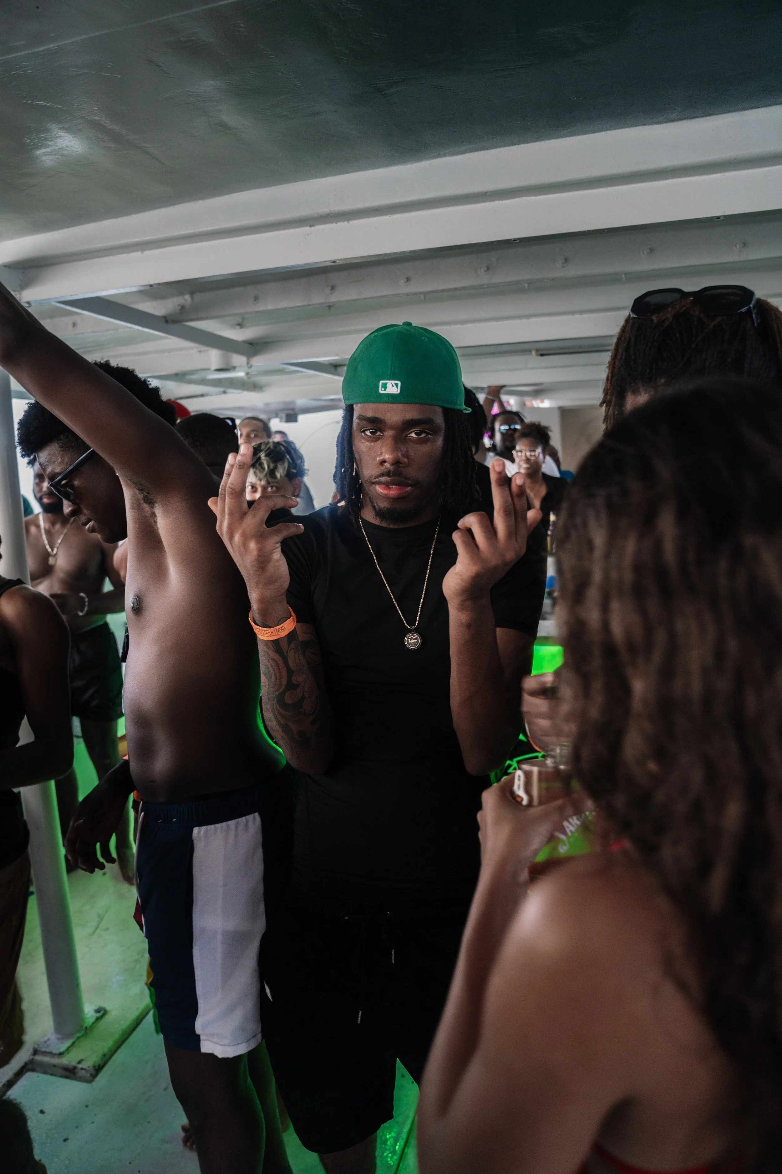 Group of young people enjoying a party on a boat, with one man wearing a green cap and making hand gestures.