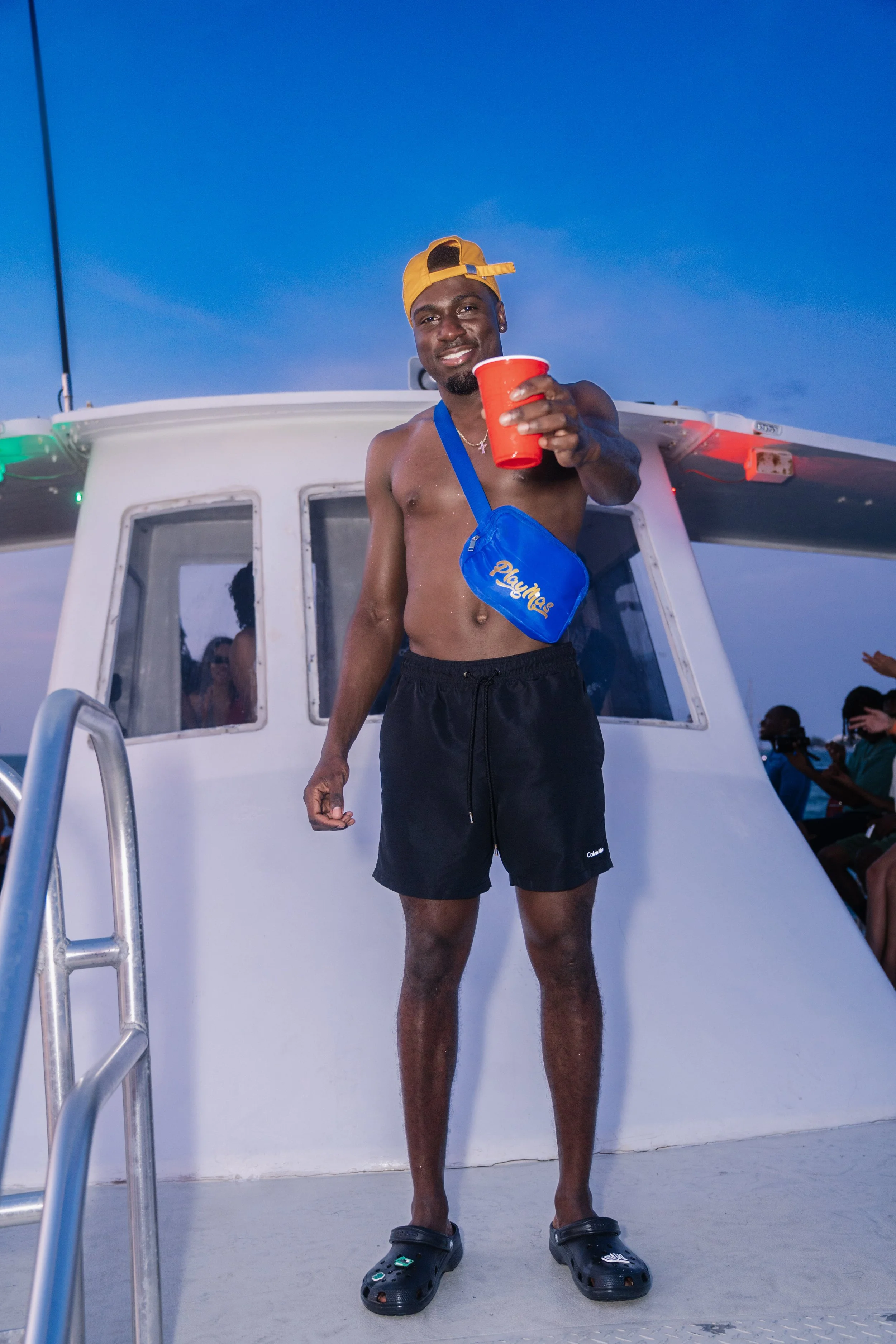 A man standing on the deck of a boat at dusk, wearing black shorts, black Crocs, a yellow cap backwards, and a blue crossbody bag, holding a red cup and smiling.