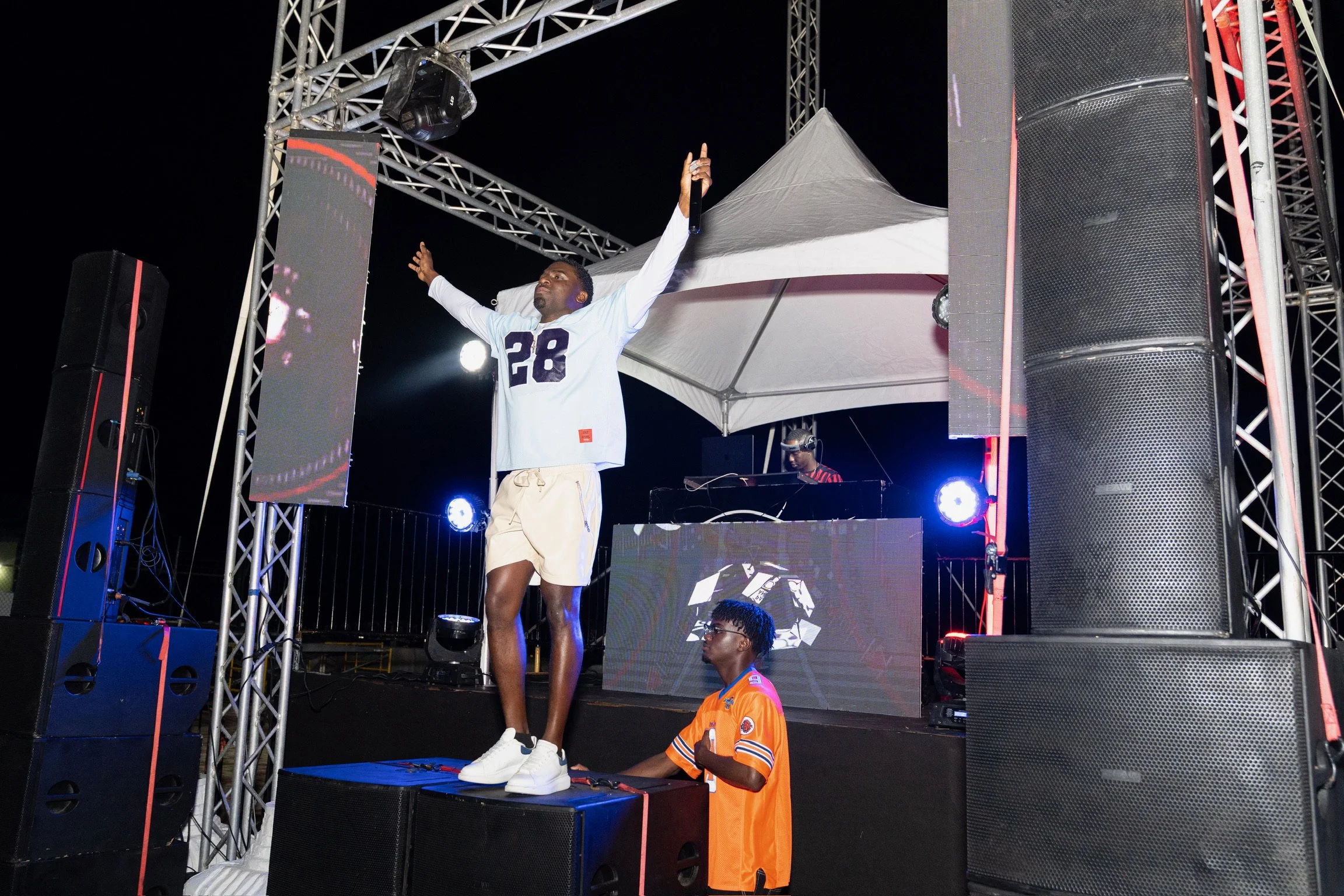 Man standing on stage with arms raised, holding a microphone, next to a DJ at a mixing console, with large speakers and lighting equipment around at night.