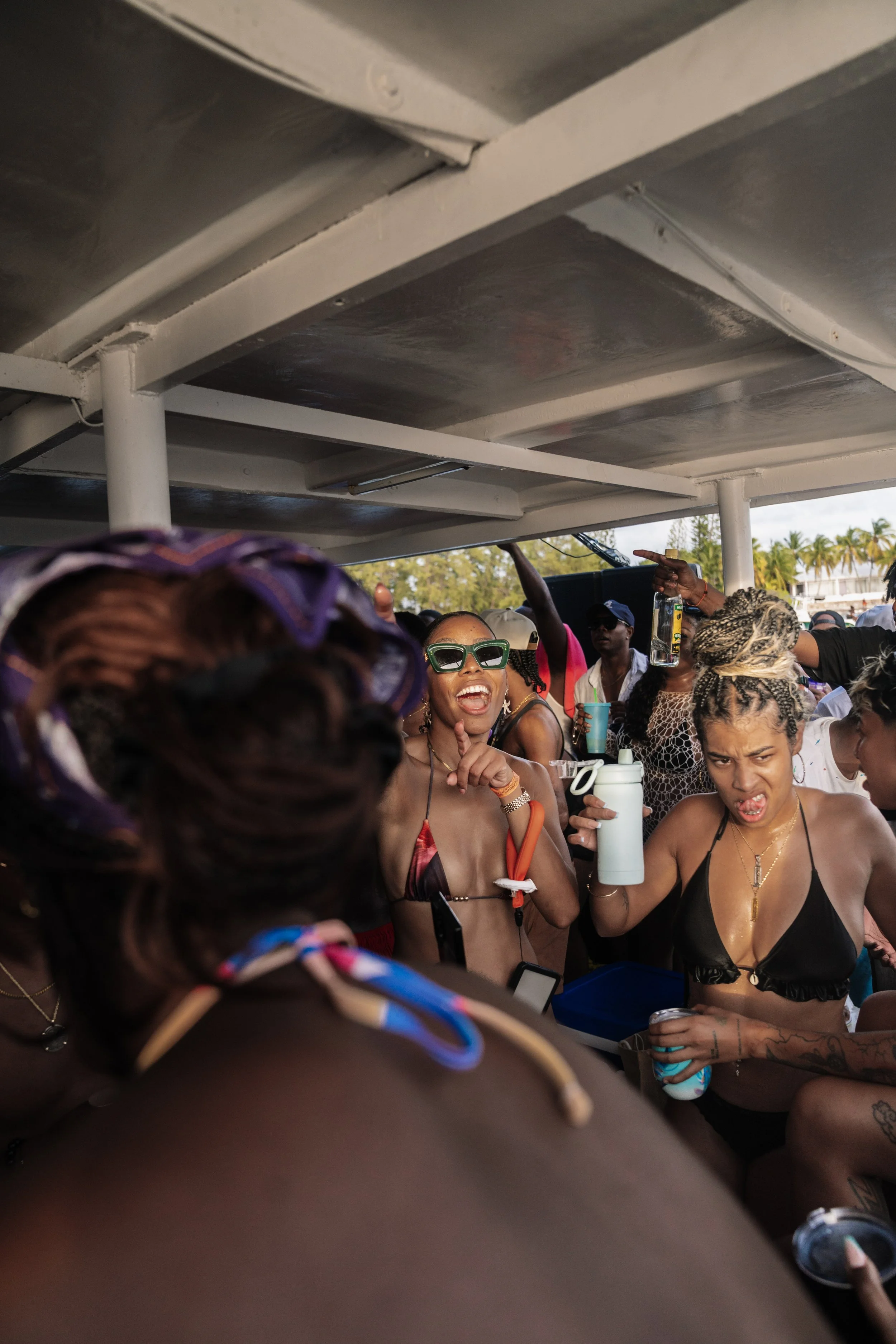 Group of people enjoying a party outdoors, with women in swimsuits dancing and smiling under a canopy, surrounded by palm trees.