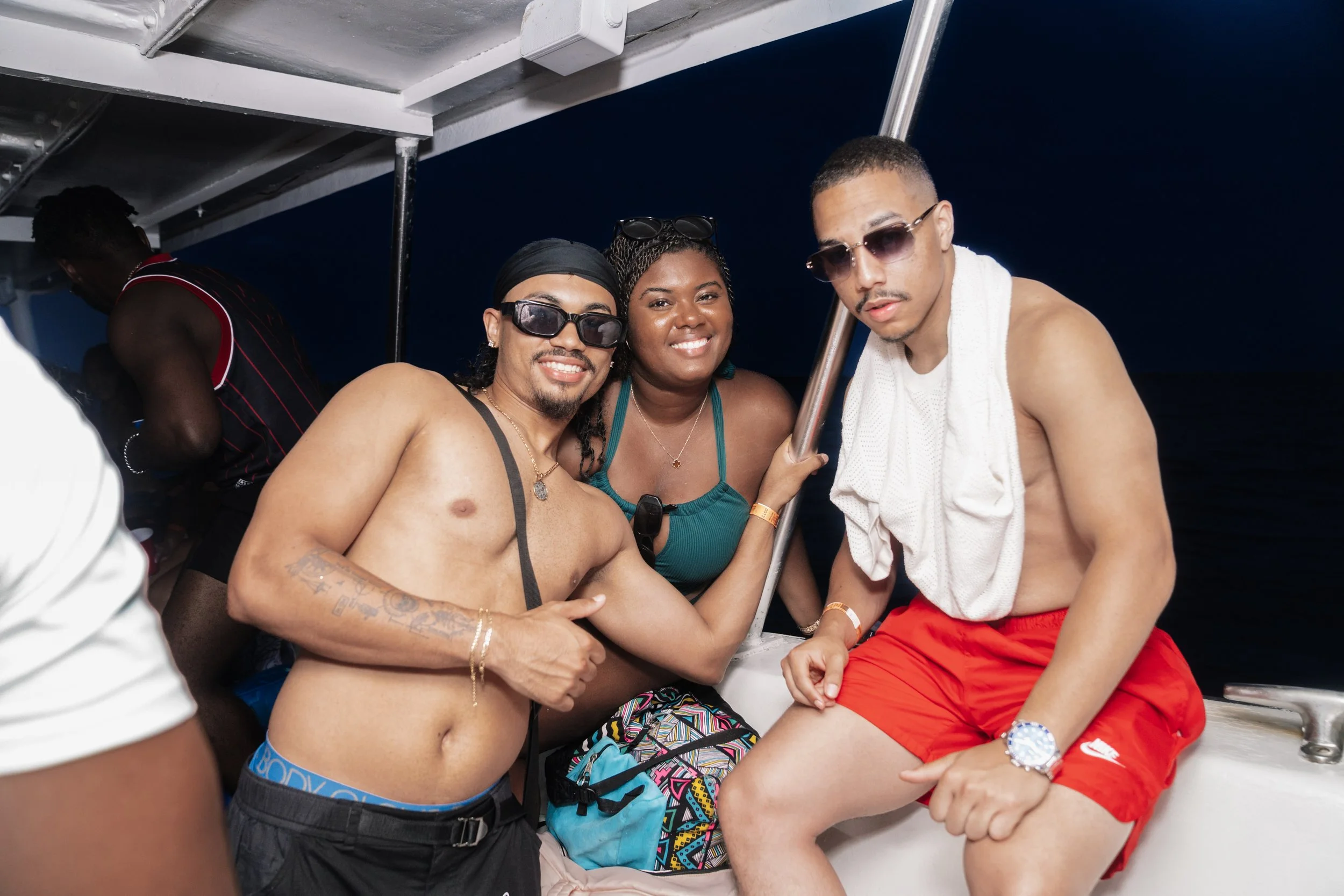 Three young adults enjoying a boat ride at night, dressed in casual summer clothing, with one shirtless man wearing sunglasses, another woman in a teal top, and a man in red shorts with a towel.