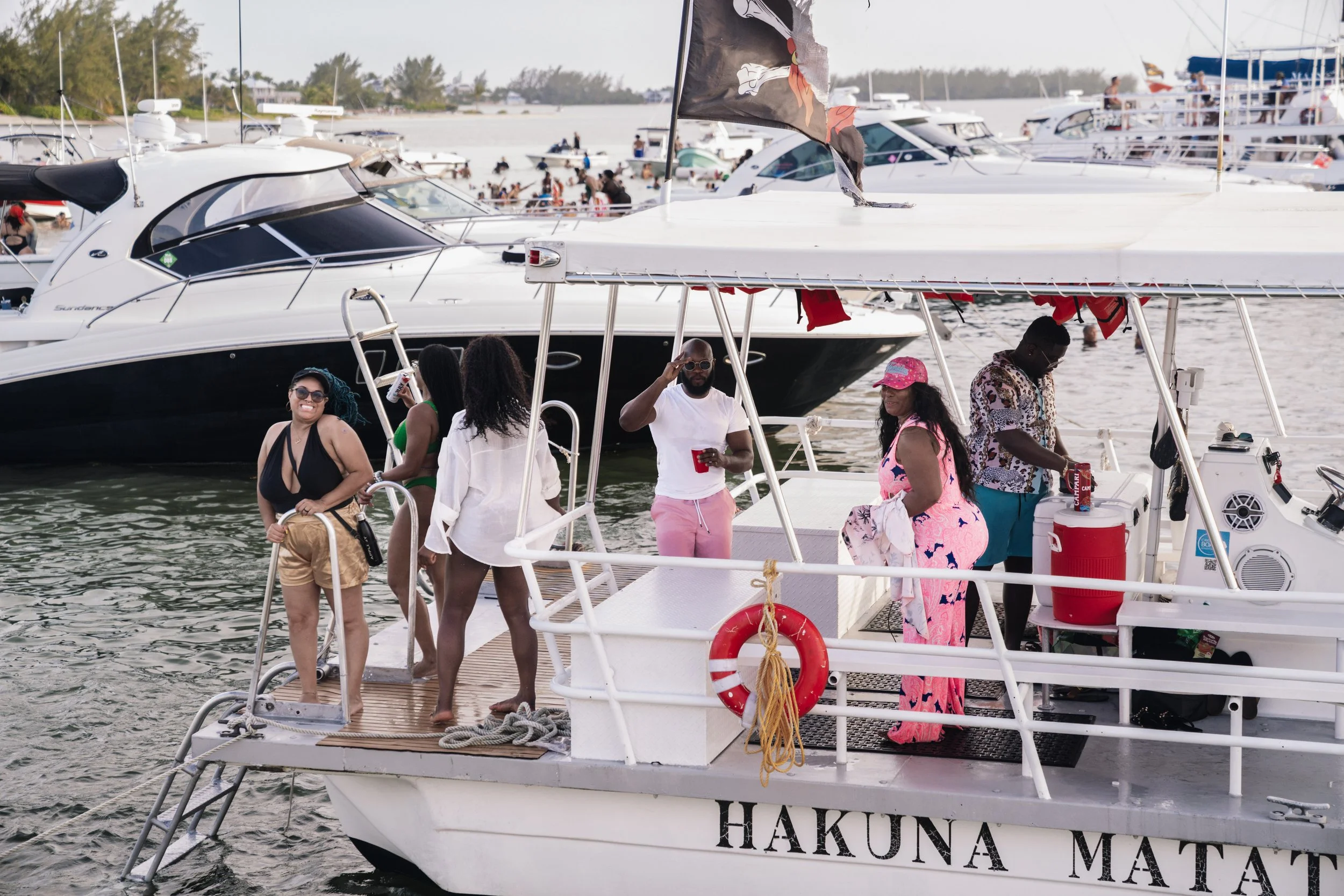 Group of people enjoying a boat party on a yacht named 'Hakuna Matata' docked at a marina, with other boats and beachgoers in the background.
