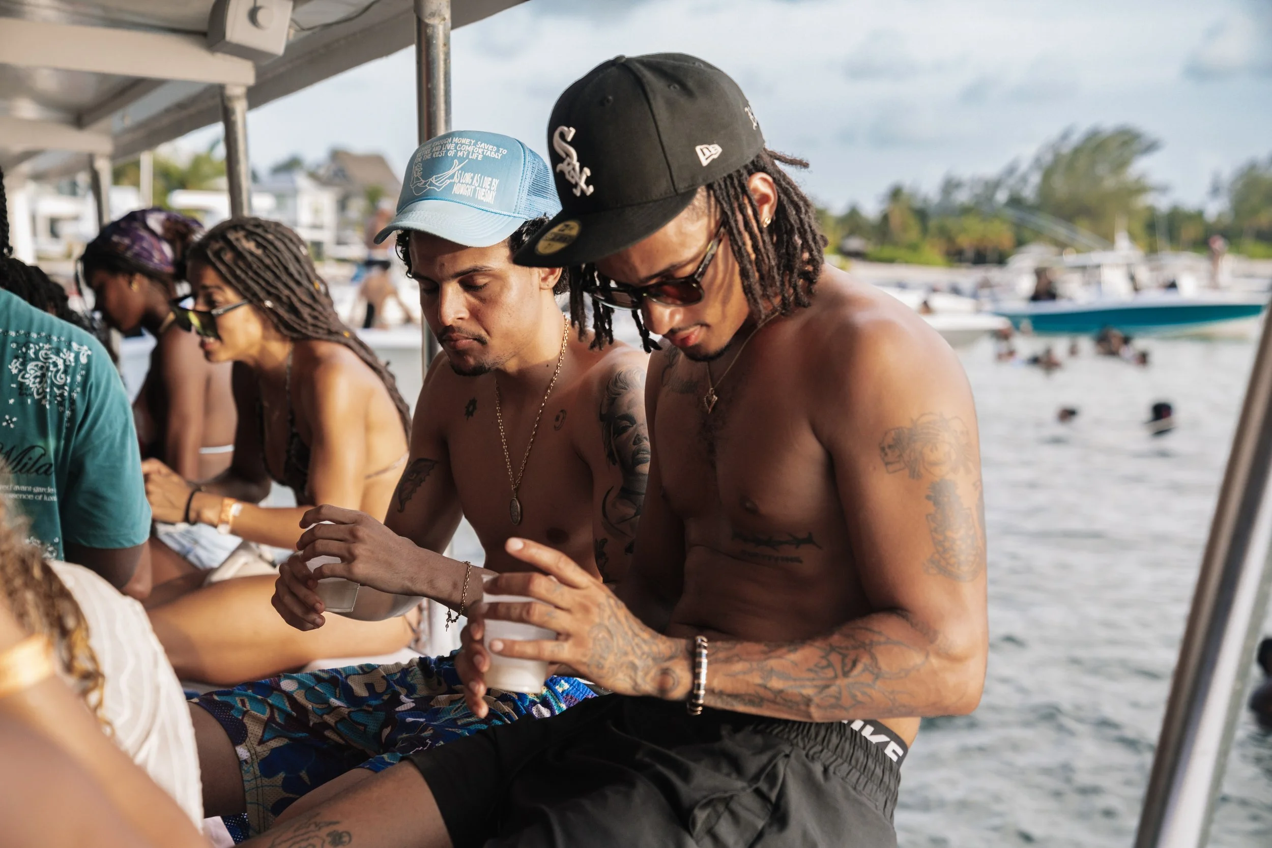 A group of shirtless men with tattoos, wearing caps and sunglasses, sitting on a boat or dock near the water holding drinks. In the background, there are boats and people swimming in a lake or river.
