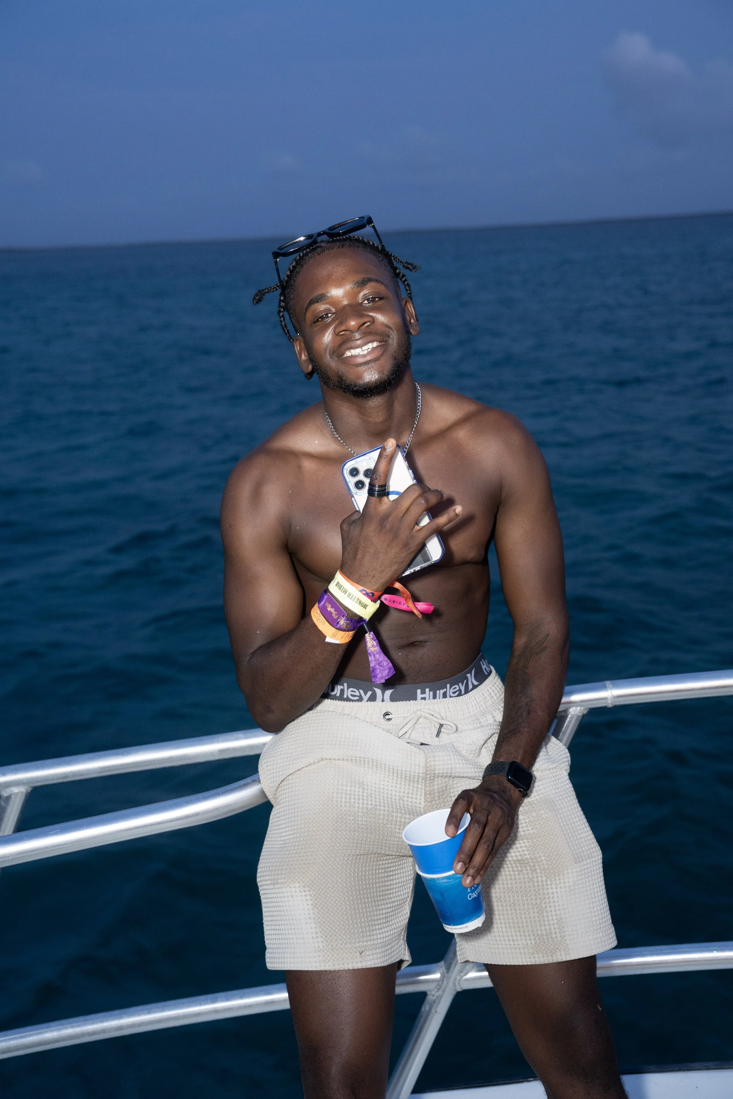 A young man with dark skin, wearing beige shorts and sunglasses on his head, holds a phone and drinks from a blue cup while standing shirtless on a boat with the ocean in the background.