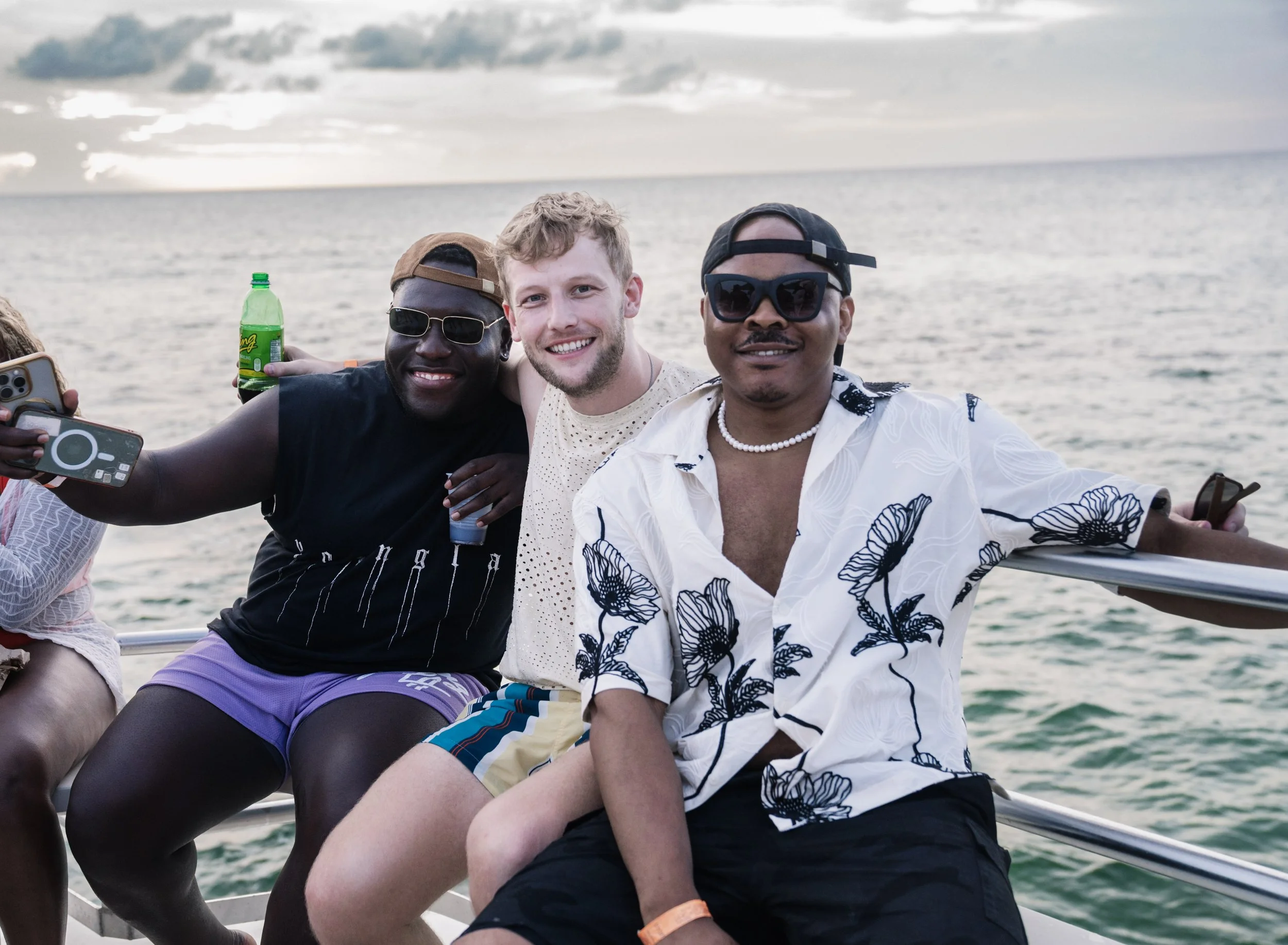 Three men sitting on a boat rail with the ocean and sky in the background, smiling and enjoying a sunny day.
