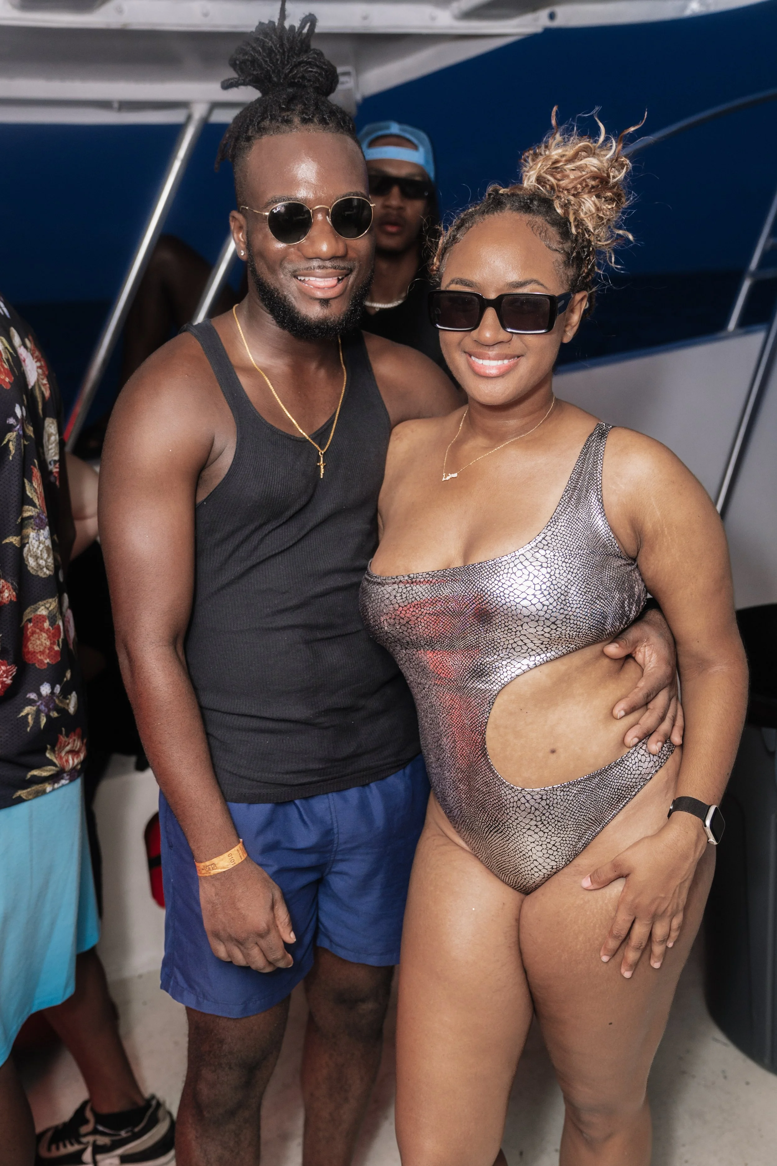 A man and woman in swimsuits smiling together at a boat party, both wearing sunglasses and standing close with arm around each other.