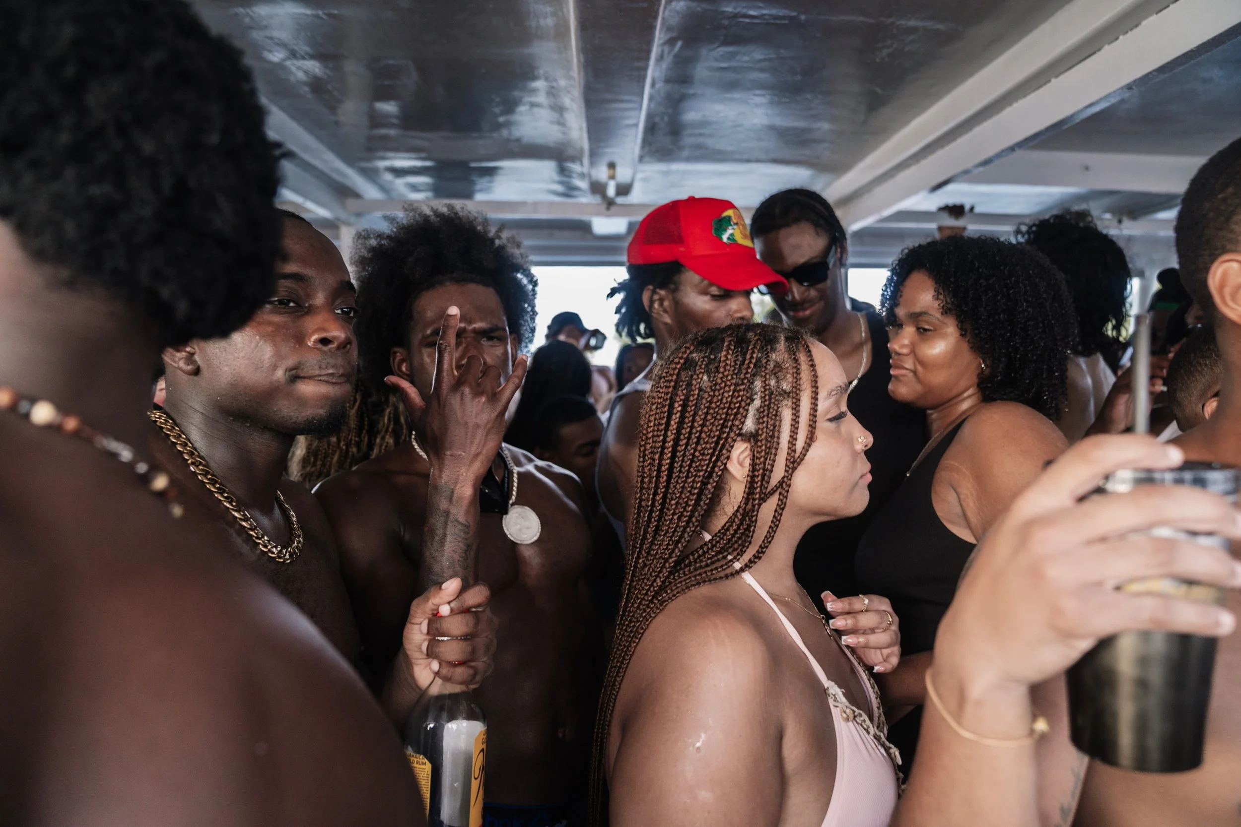 A group of people gathered on a boat, some are shirtless and wearing jewelry, with one person holding a drink and another taking a photo with a phone, enjoying a social event.