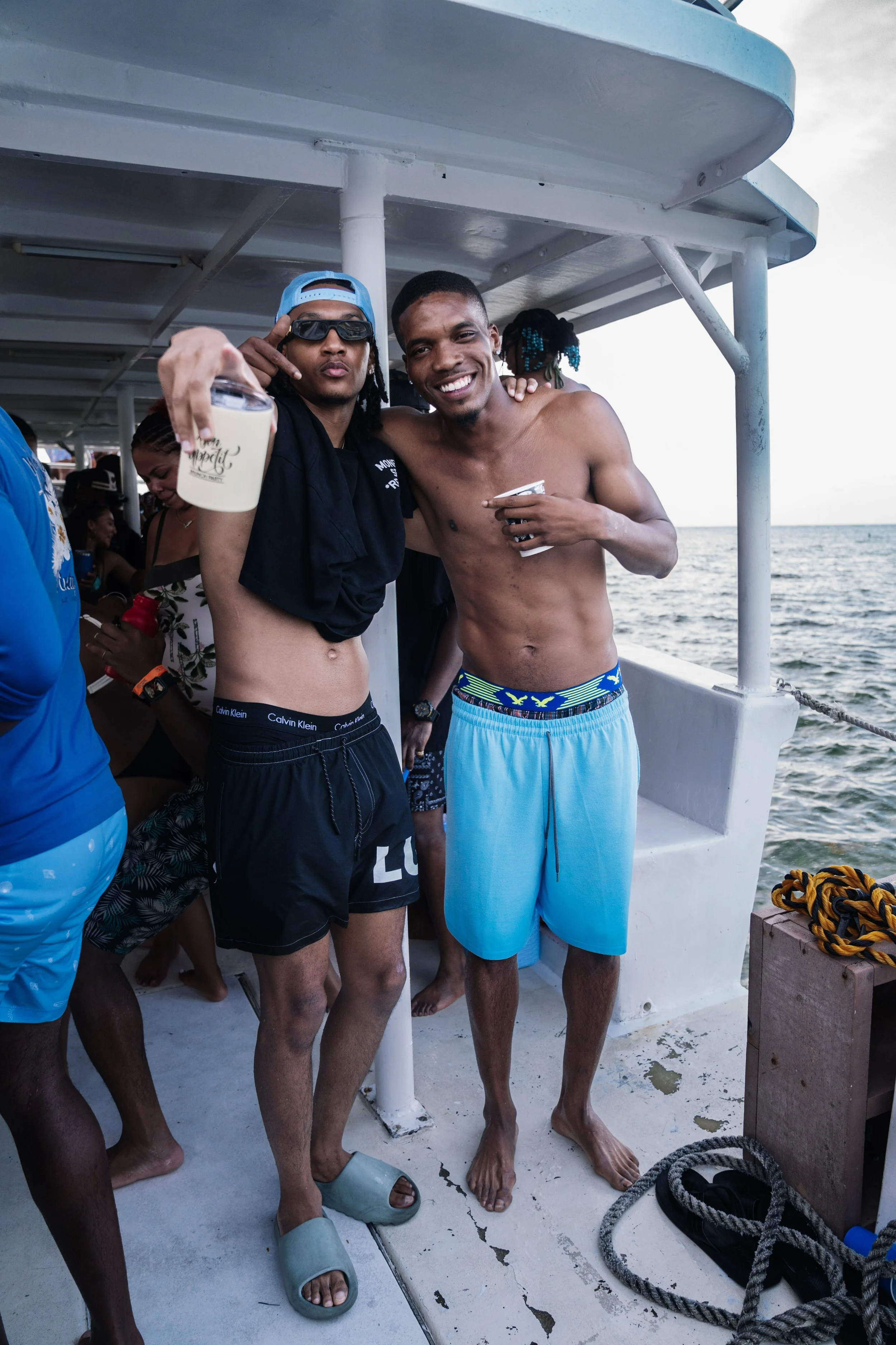 Two young men in swim trunks smiling and posing on a boat, with the ocean and other people in the background.