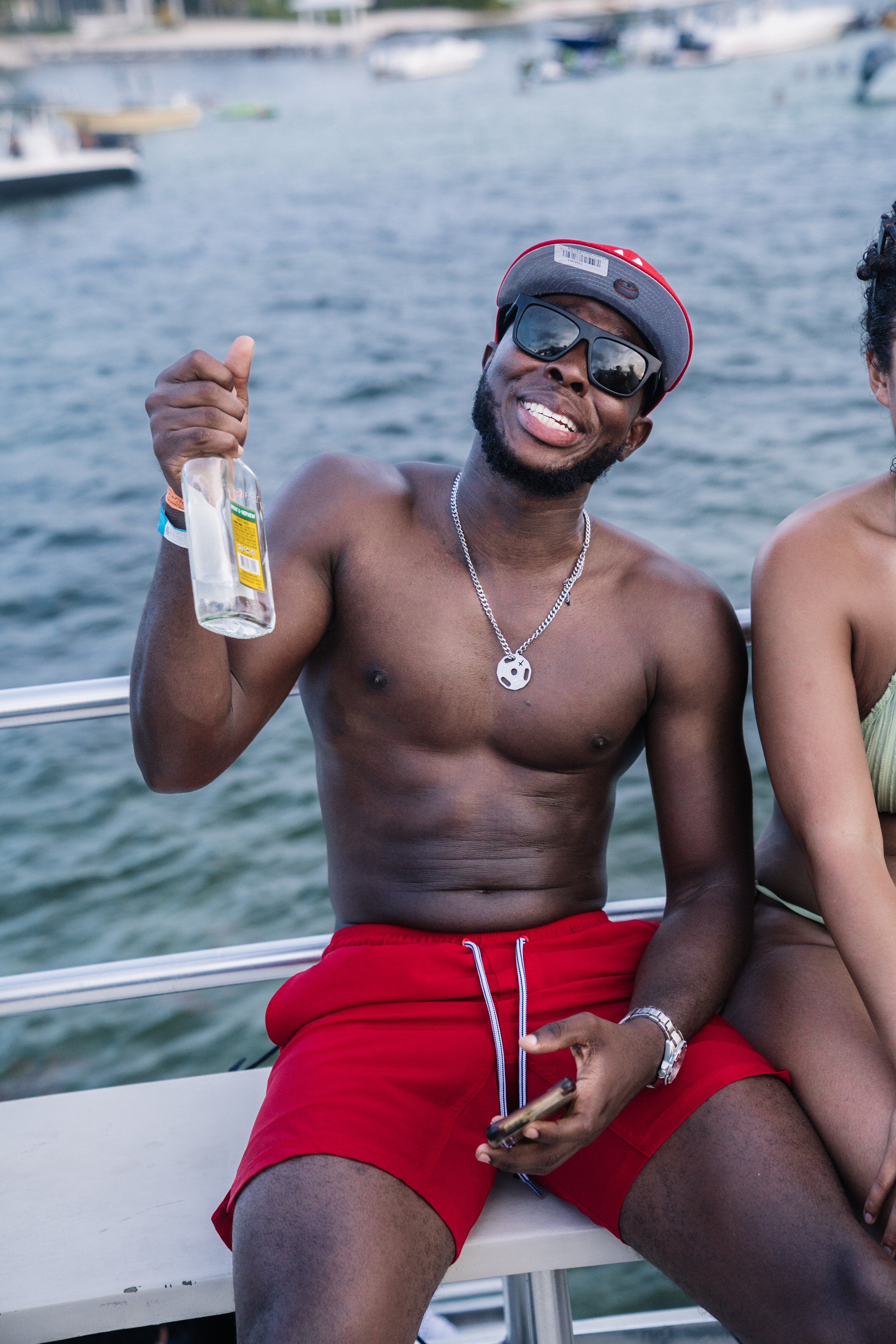 A shirtless young man with sunglasses, a cap, and a necklace, sitting on a boat, holding a bottle, smiling, with a lake and boats in the background.