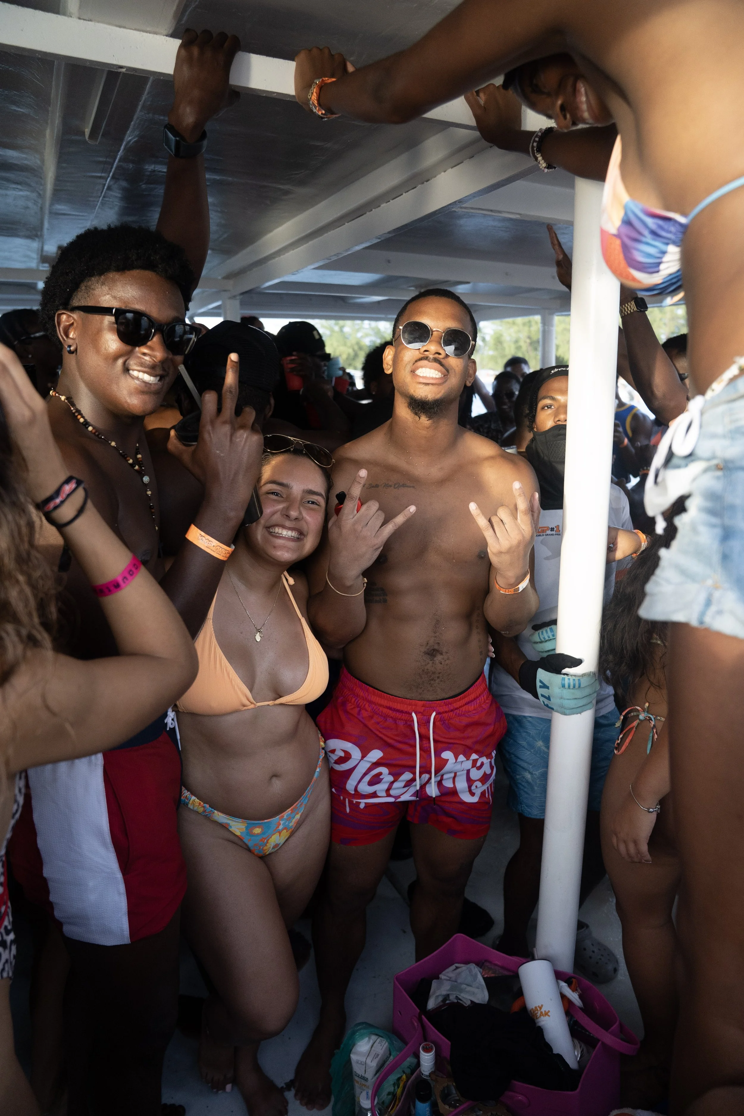 Group of young adults enjoying a boat party on a sunny day, wearing swimsuits and sunglasses, with some smiling and making hand gestures.