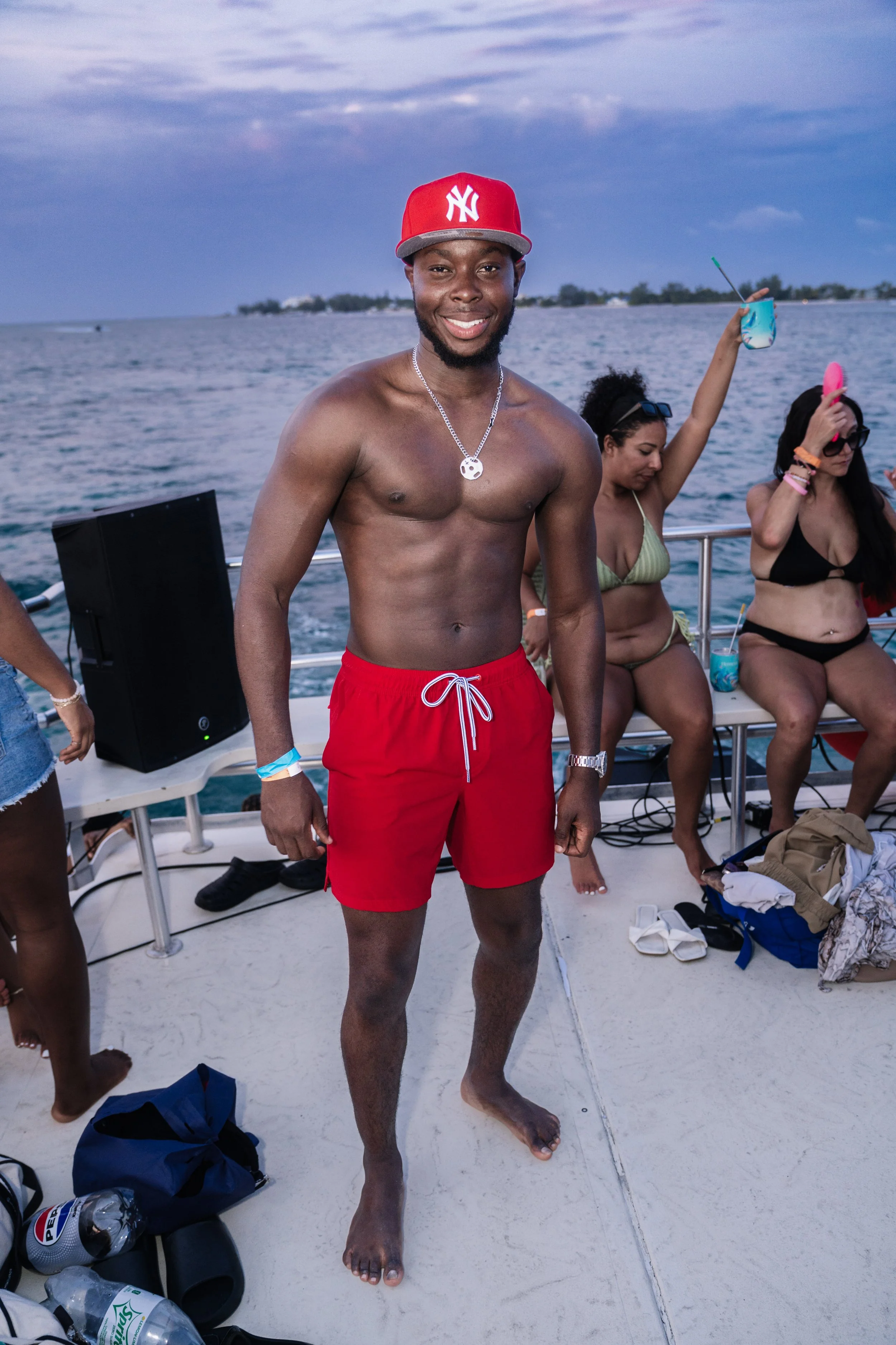 A young man smiling and posing on a boat in a red New York Yankees cap and red swim trunks, with two women in bikinis sitting behind him, surrounded by water and a partly cloudy sky during daytime.