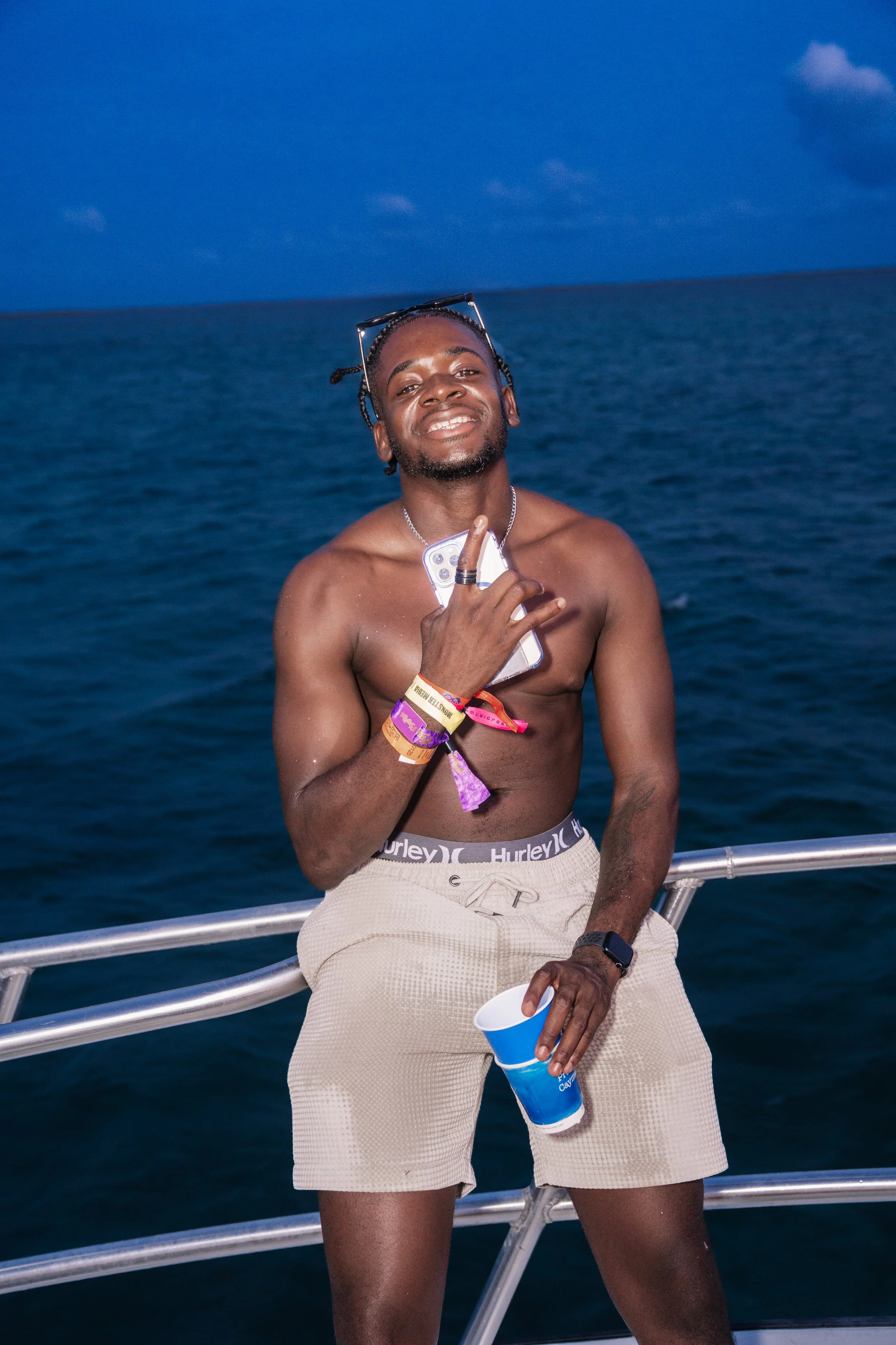 A shirtless man with glasses on his head, wearing beige shorts and numerous wristbands, holding a cup in one hand and a cellphone in the other, standing on a boat with a body of water and cloudy sky in the background.