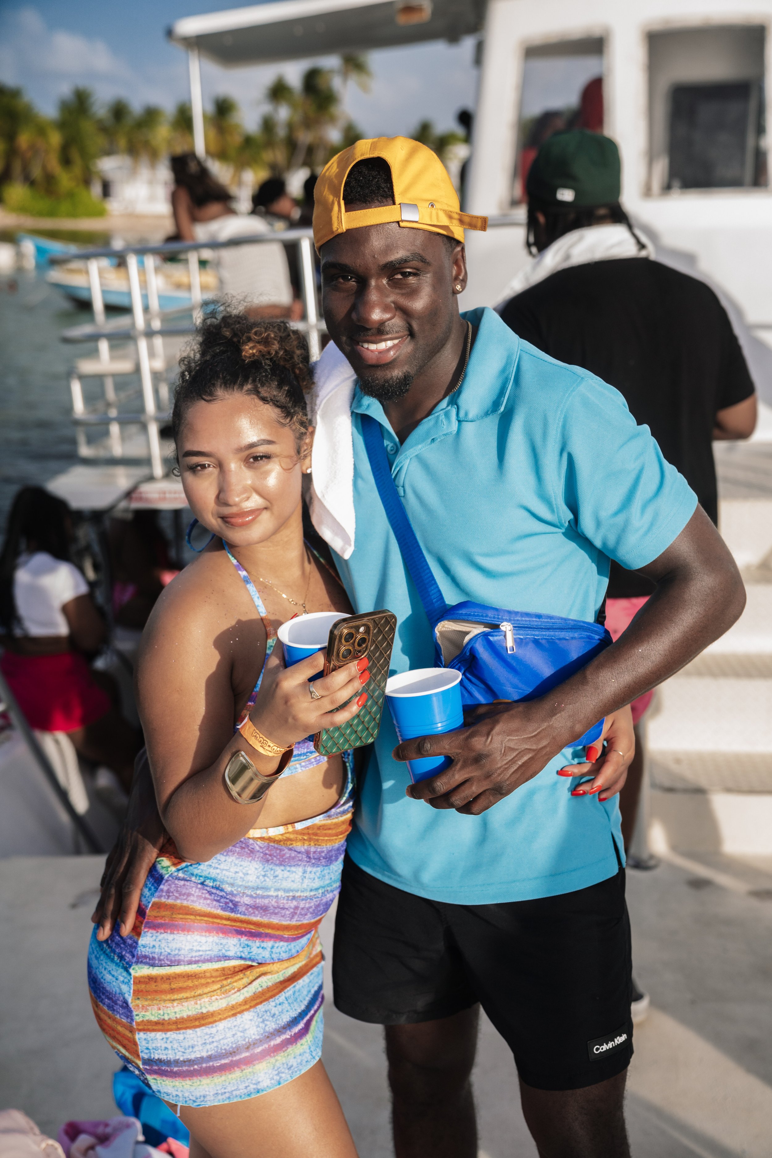 Two people posing on a boat deck, smiling, with a woman holding a phone and a man holding cups, wearing casual summer clothing, with a boat and water in the background.
