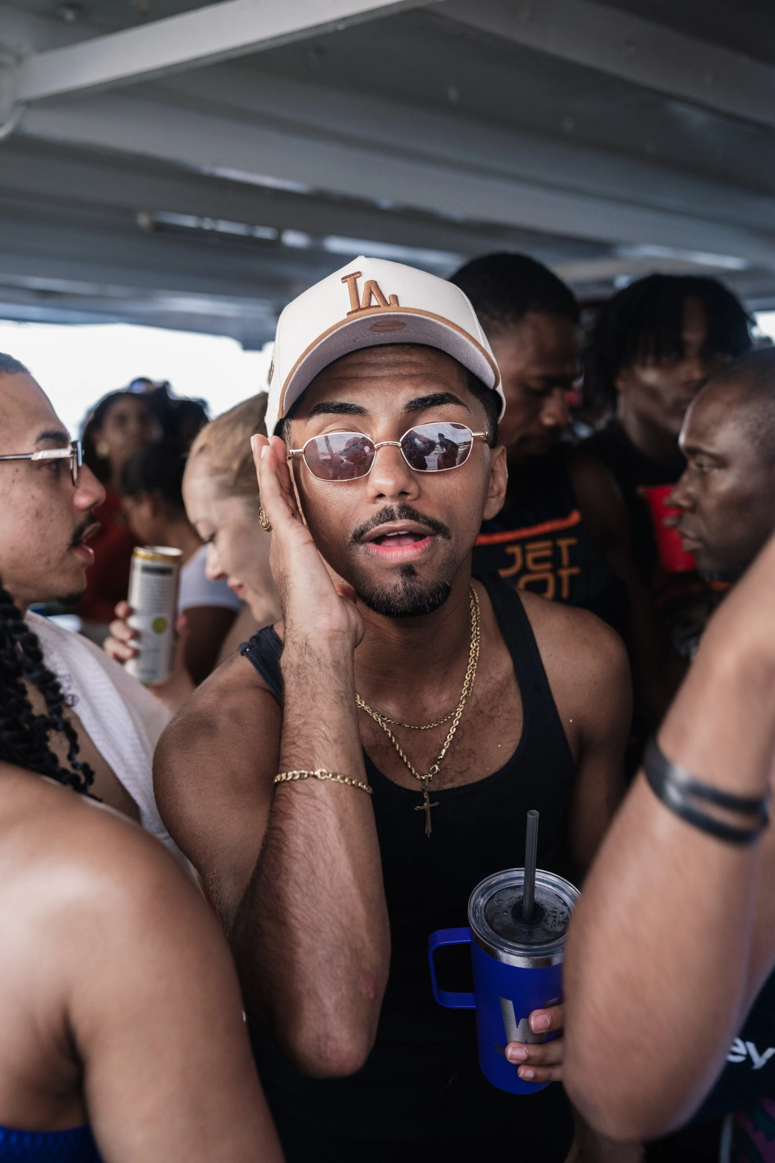 A man wearing sunglasses, a white baseball cap with 'LA' on it, and gold jewelry poses for a photo among a crowd at a party or event, holding a drink in a blue container.