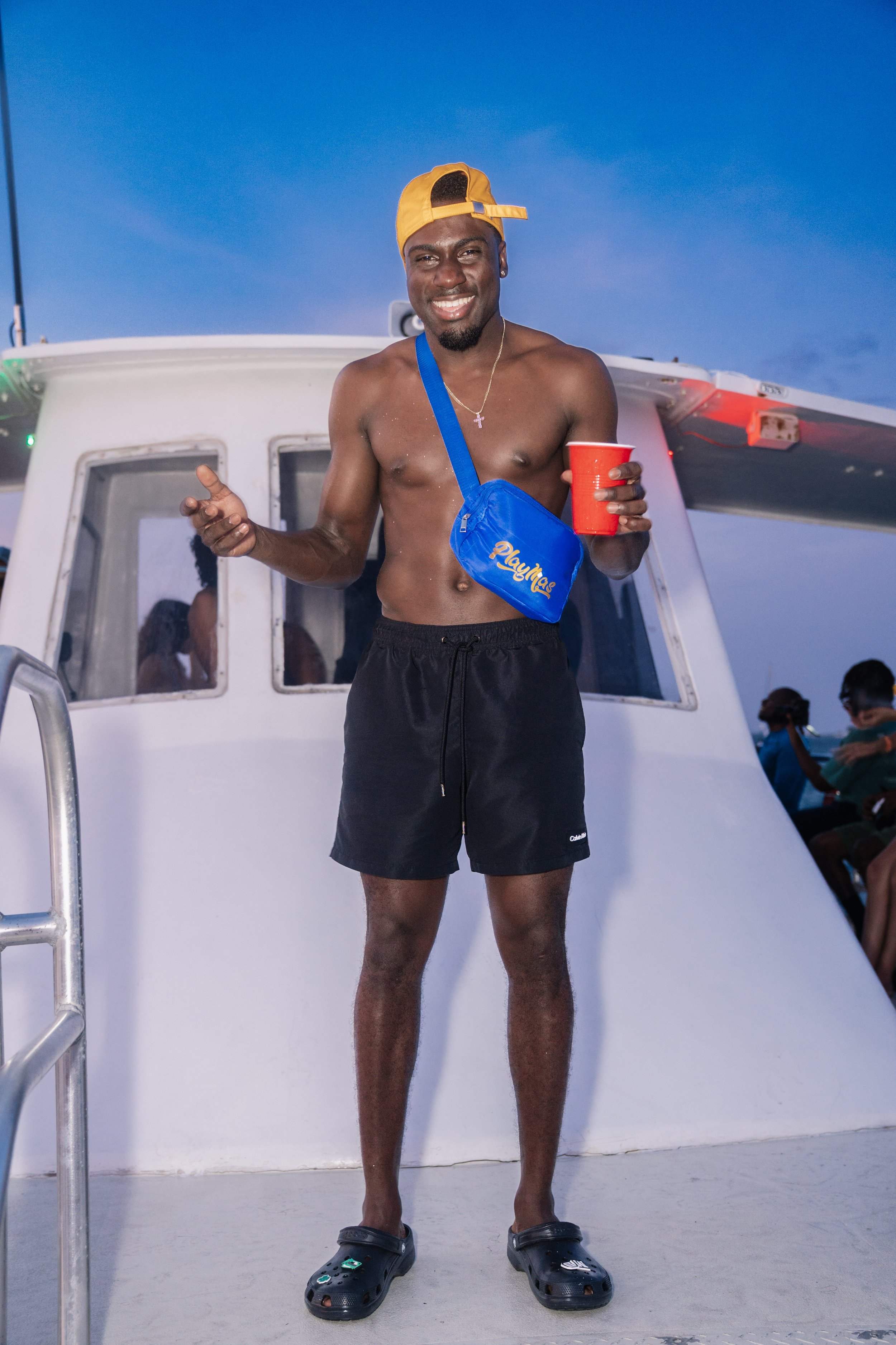 A shirtless man wearing a yellow baseball cap, black shorts, and black Crocs shoes standing on a boat at dusk, holding red cup in one hand and a blue crossbody bag.