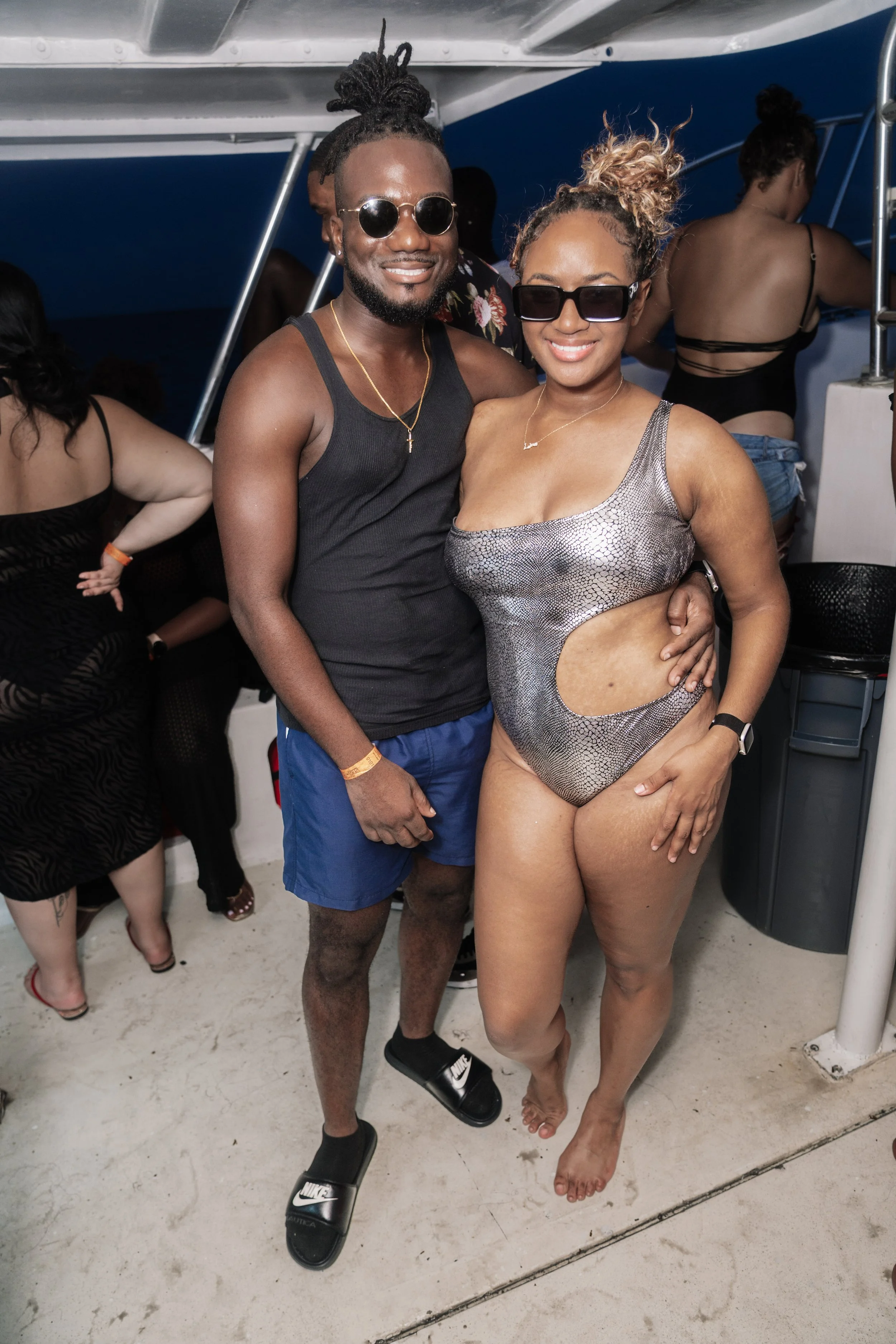 A man and woman smiling and posing together on a boat at night, with other people in swimwear in the background.