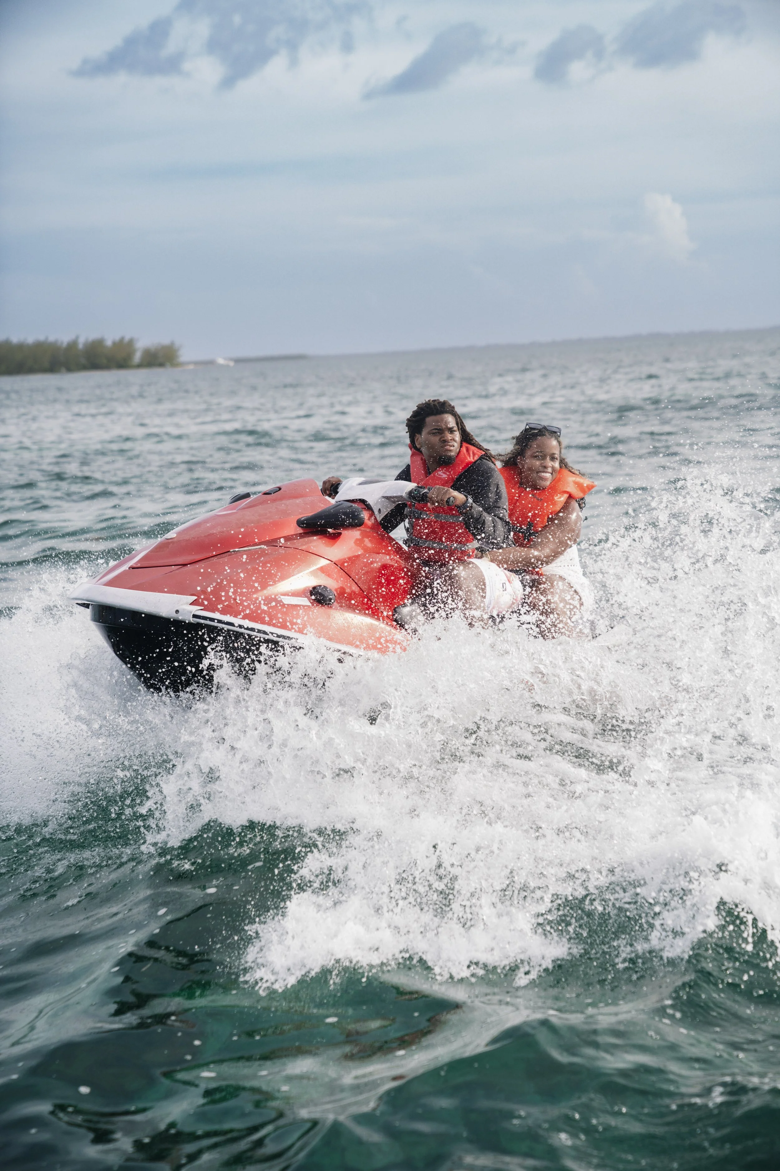 Two people riding a red jet ski on the water, both wearing orange life jackets, with a cloudy sky above and a distant shoreline.