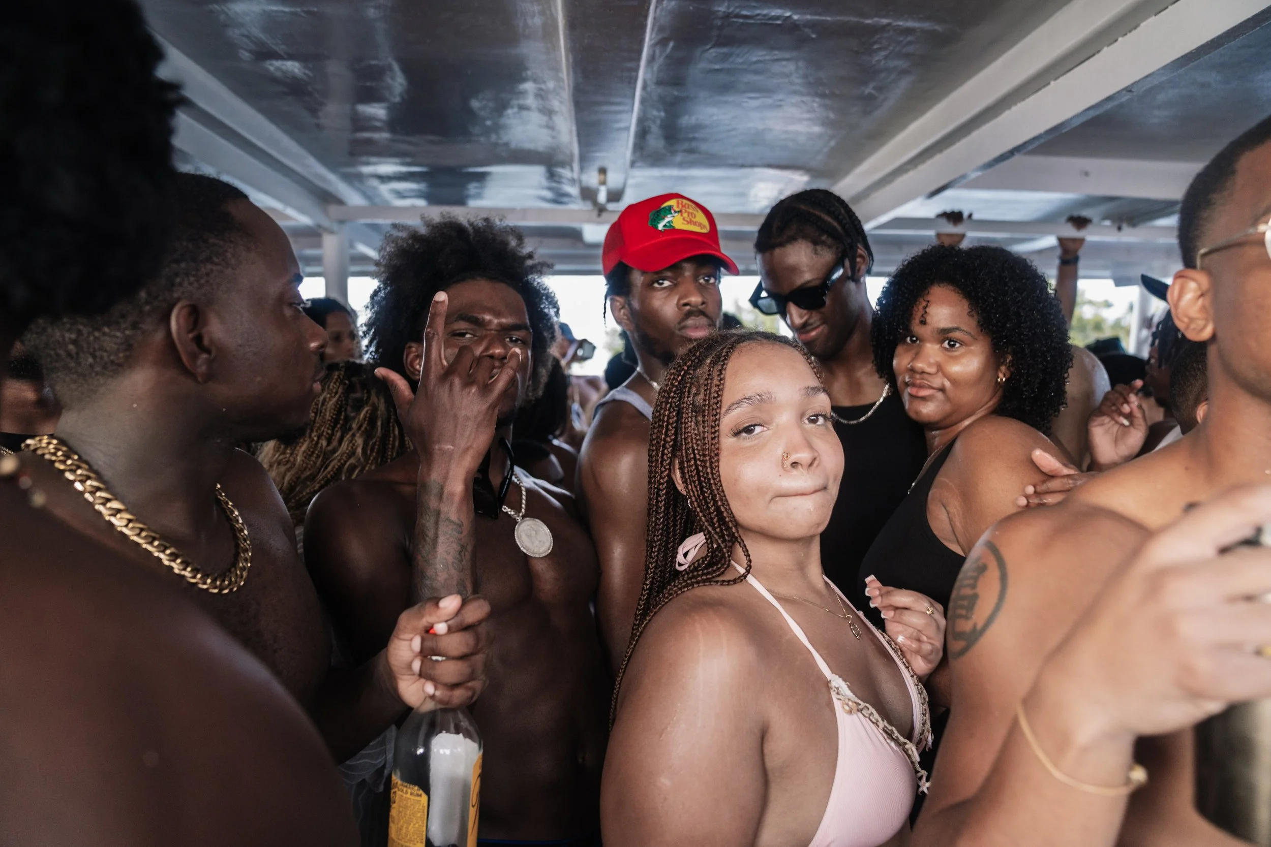 Group of young adults at a party or social gathering, some shirtless, wearing casual clothes and accessories, under a shaded area, with one person holding a bottle of alcohol.