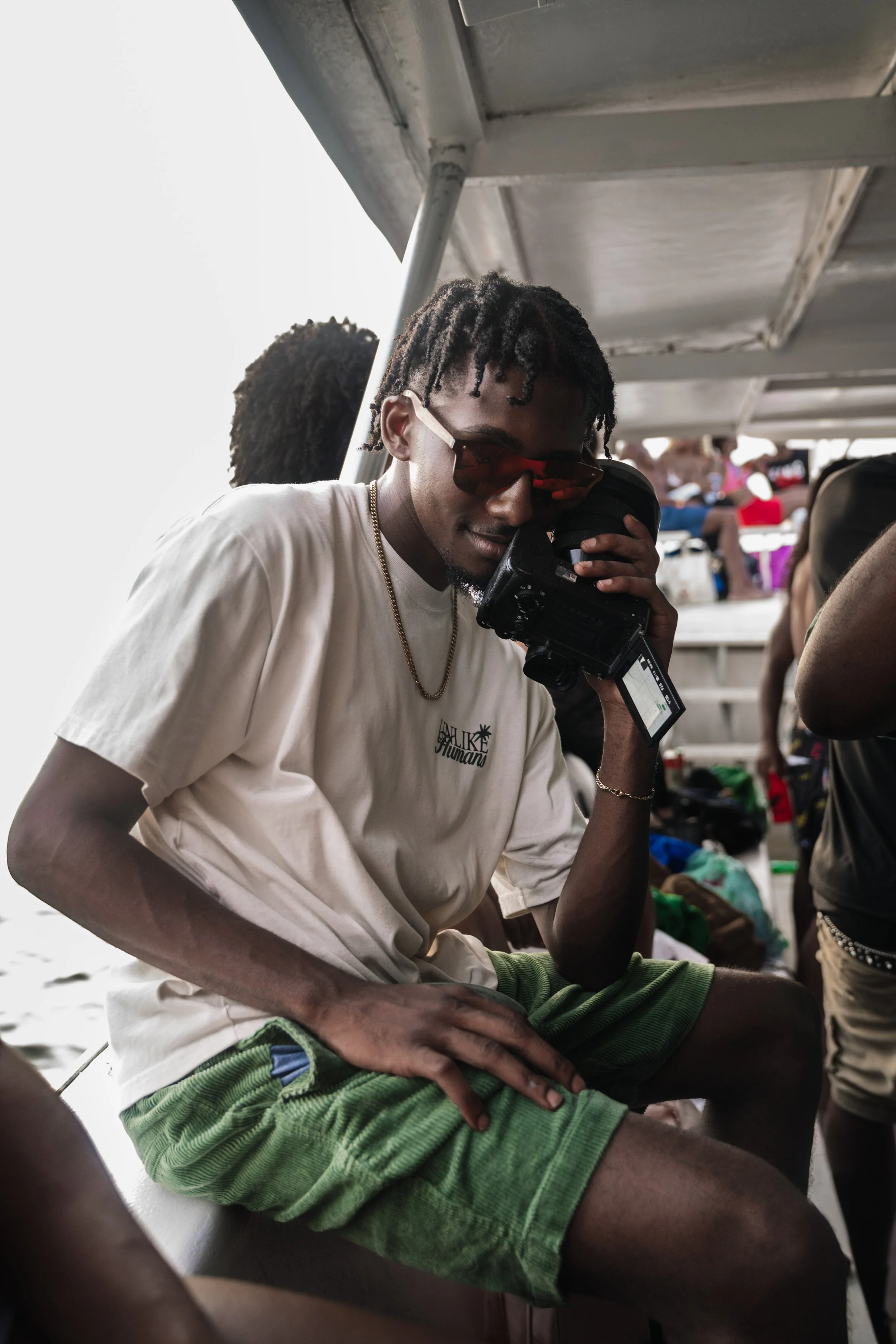 Young man with sunglasses filming with a camera on a boat with people in the background.