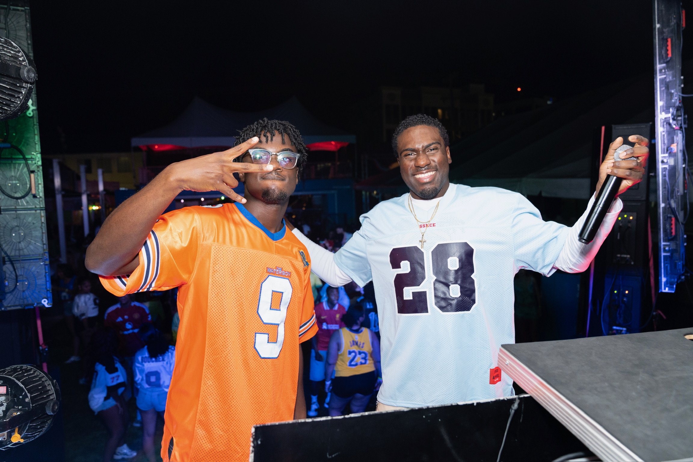 Two men at a nighttime event; one in an orange sports jersey making a peace sign, the other in a white jersey holding a microphone, smiling, DJ equipment in the foreground.