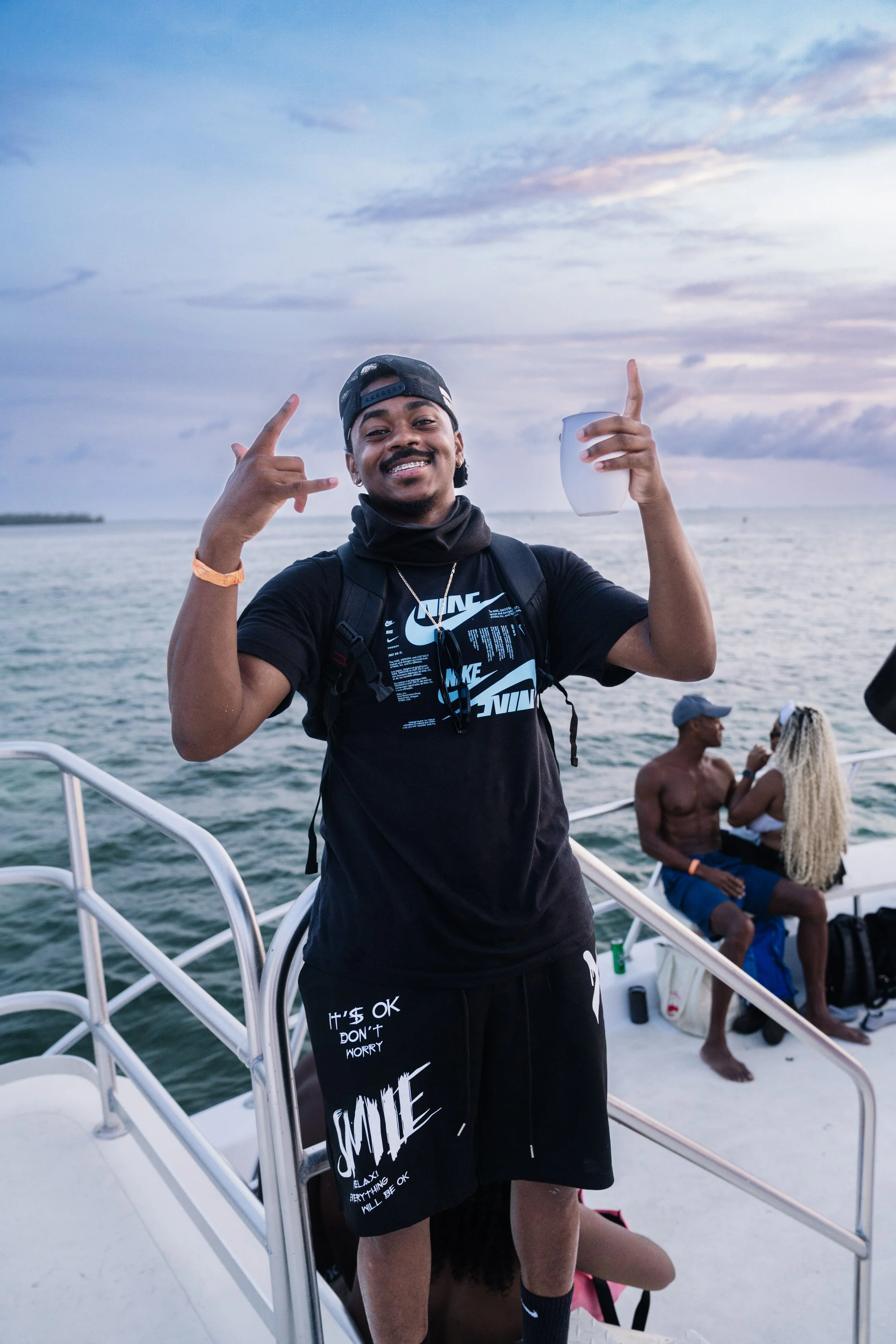A young man smiling and making rock and roll hand gestures while on a boat, holding a white cup, with two people sitting in the background near the water during sunset.