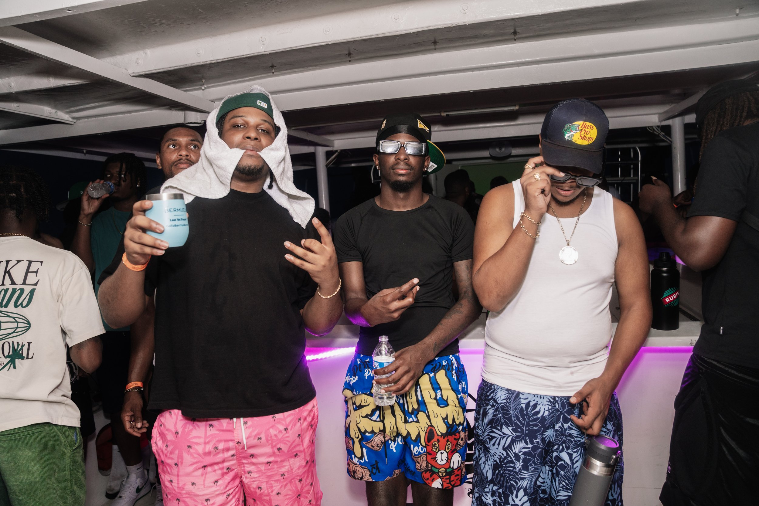 Three men at a party or nightclub. The man on the left wears a black shirt with pink patterned shorts and has a towel around his head. He is holding a drink and making a hand gesture. The man in the middle wears a black t-shirt, colorful shorts, glas
