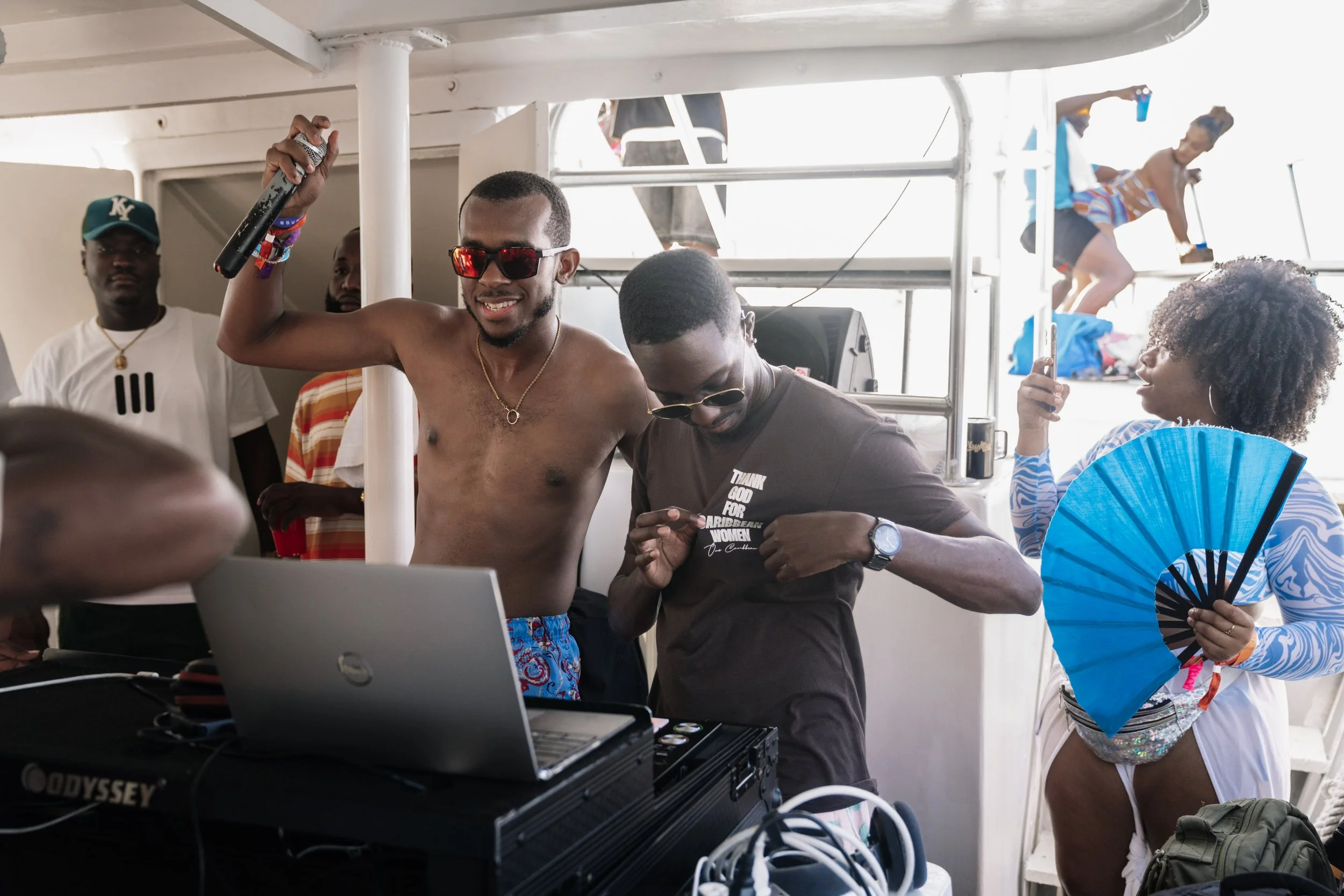 Group of people dancing and enjoying music on a boat, with a shirtless man holding a microphone, a woman with a fan, and others having fun in the background.