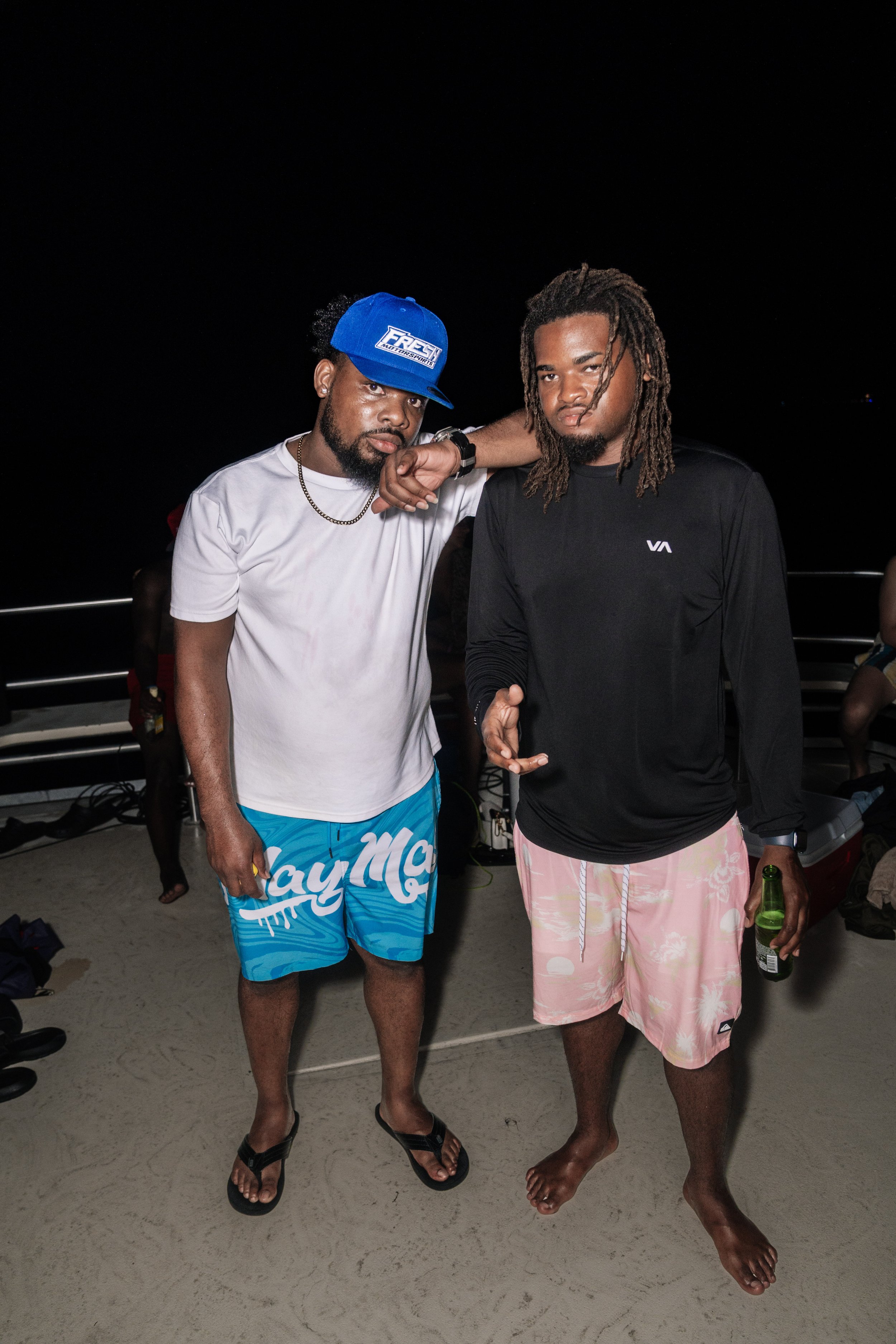 Two young men standing on a deck at night, one wearing a white T-shirt, blue shorts, a blue cap, and flip-flops, the other in a black long-sleeve shirt and pink swim trunks, holding a beer, with a dark sky and some people in the background.