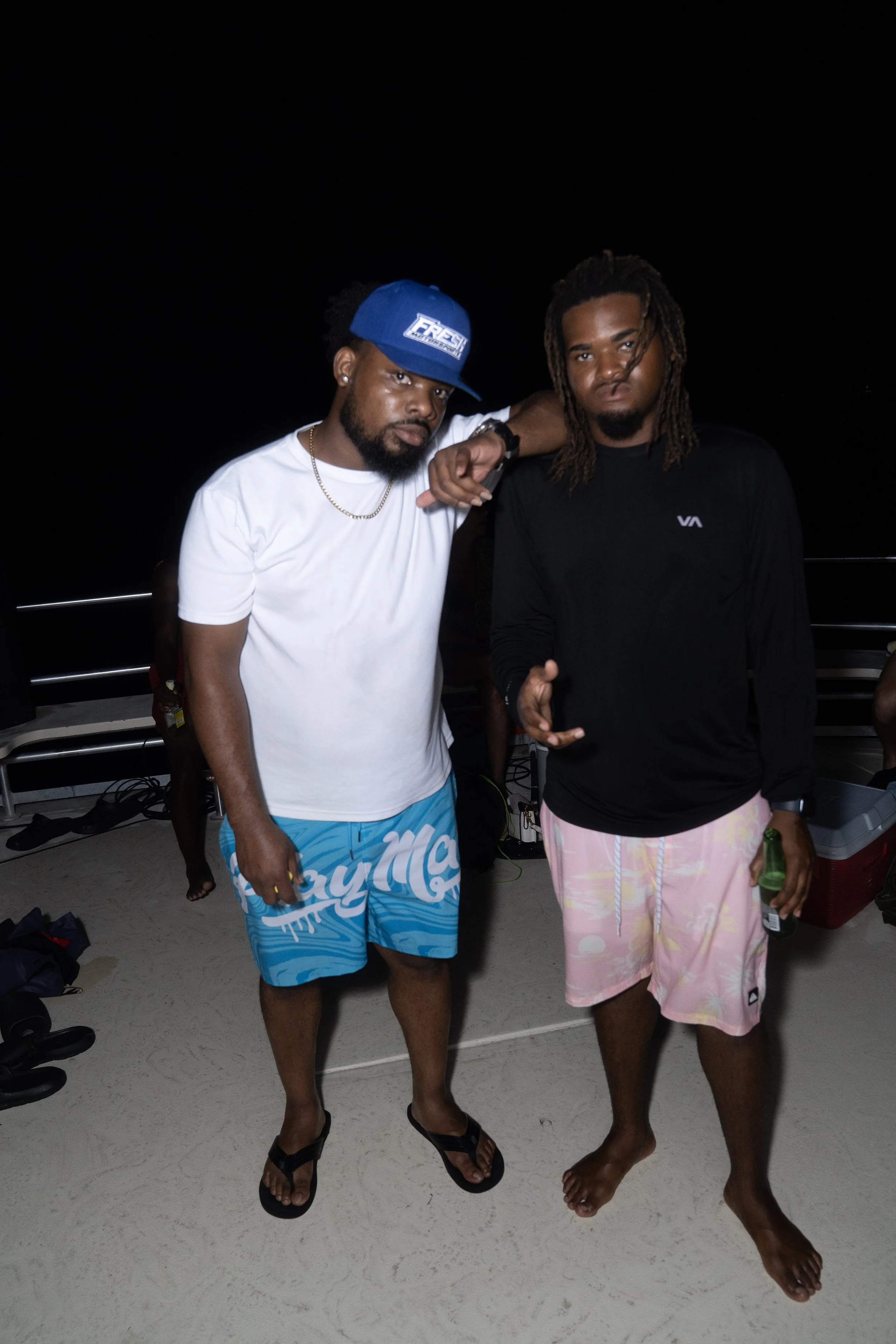 Two young men in casual beachwear posing at night on a sandy surface with a dark background.