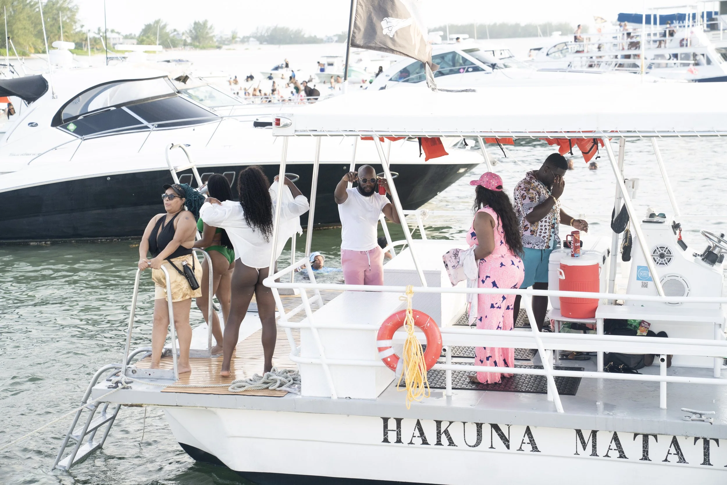 A group of people on a boat at a marina, with some swimming in the water and others socializing on the deck. The boat has the name 'Hakuna Matata'.