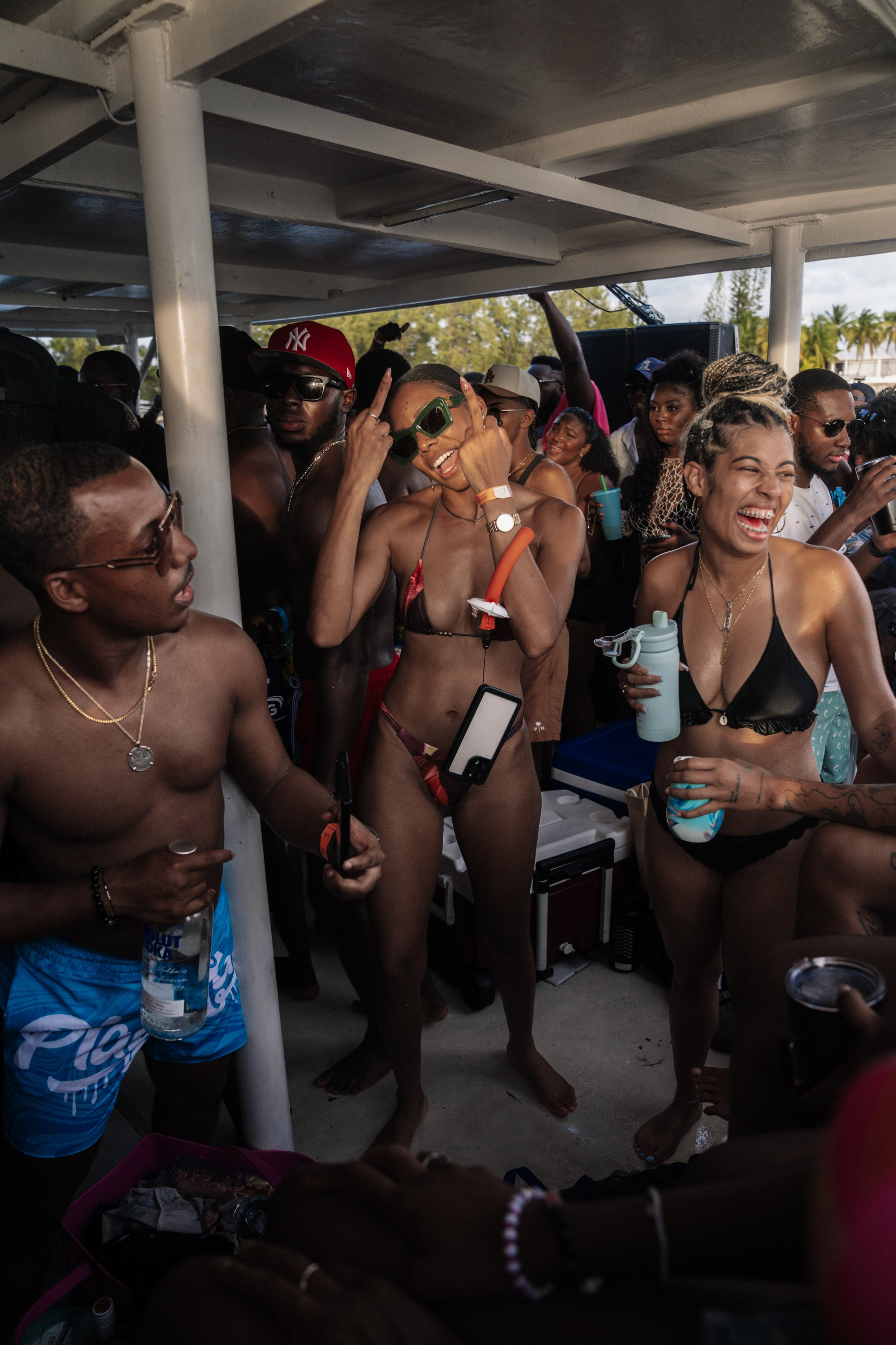 Group of young adults enjoying a pool party, some in swimsuits, drinking and dancing in a covered outdoor area with palm trees in the background.
