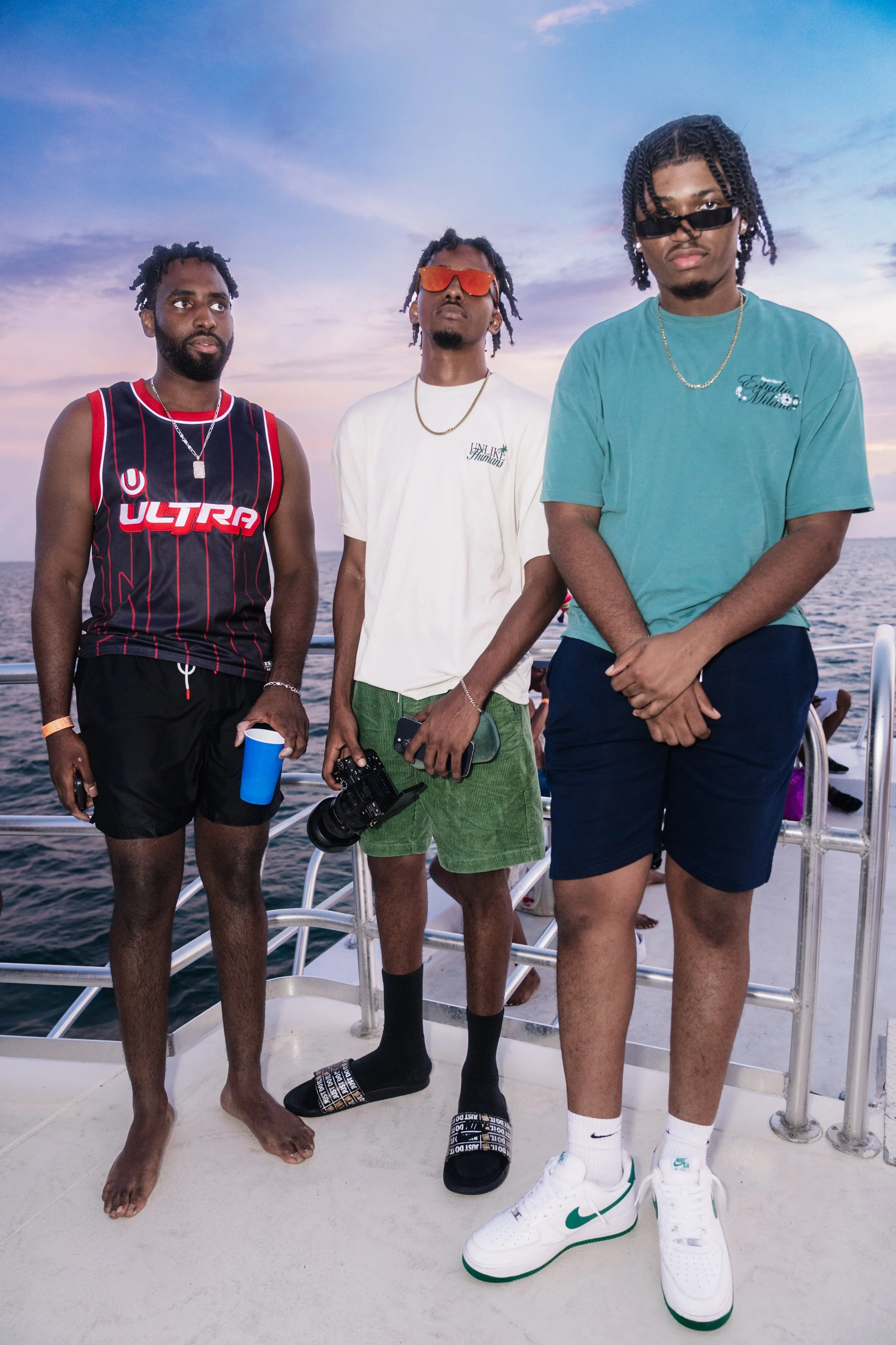 Three young men standing on a boat at sunset, dressed in casual summer clothing, with the ocean in the background.