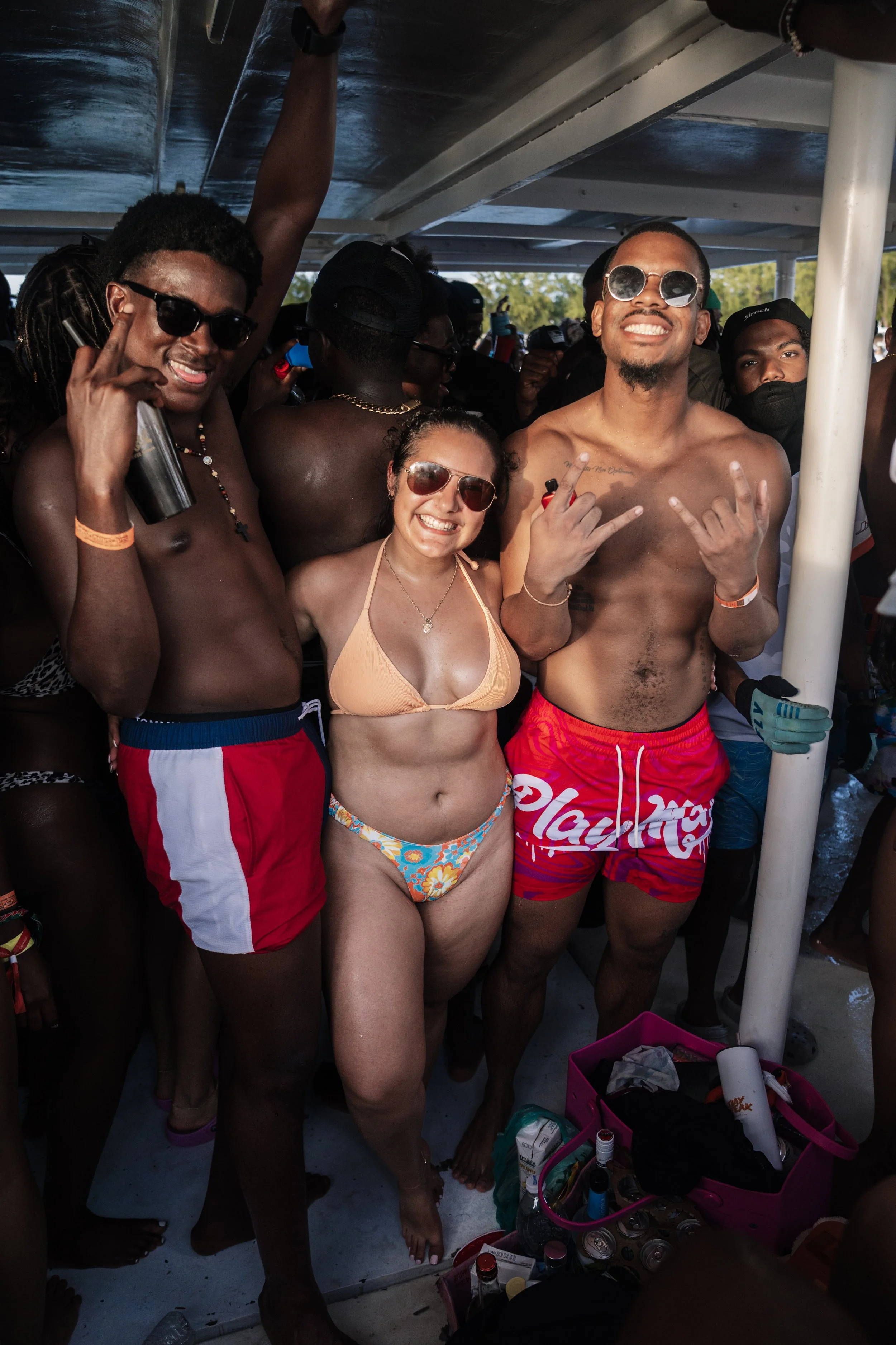 People enjoying a boat party, dressed in swimsuits, some wearing sunglasses, with drinks and a pink cooler visible.
