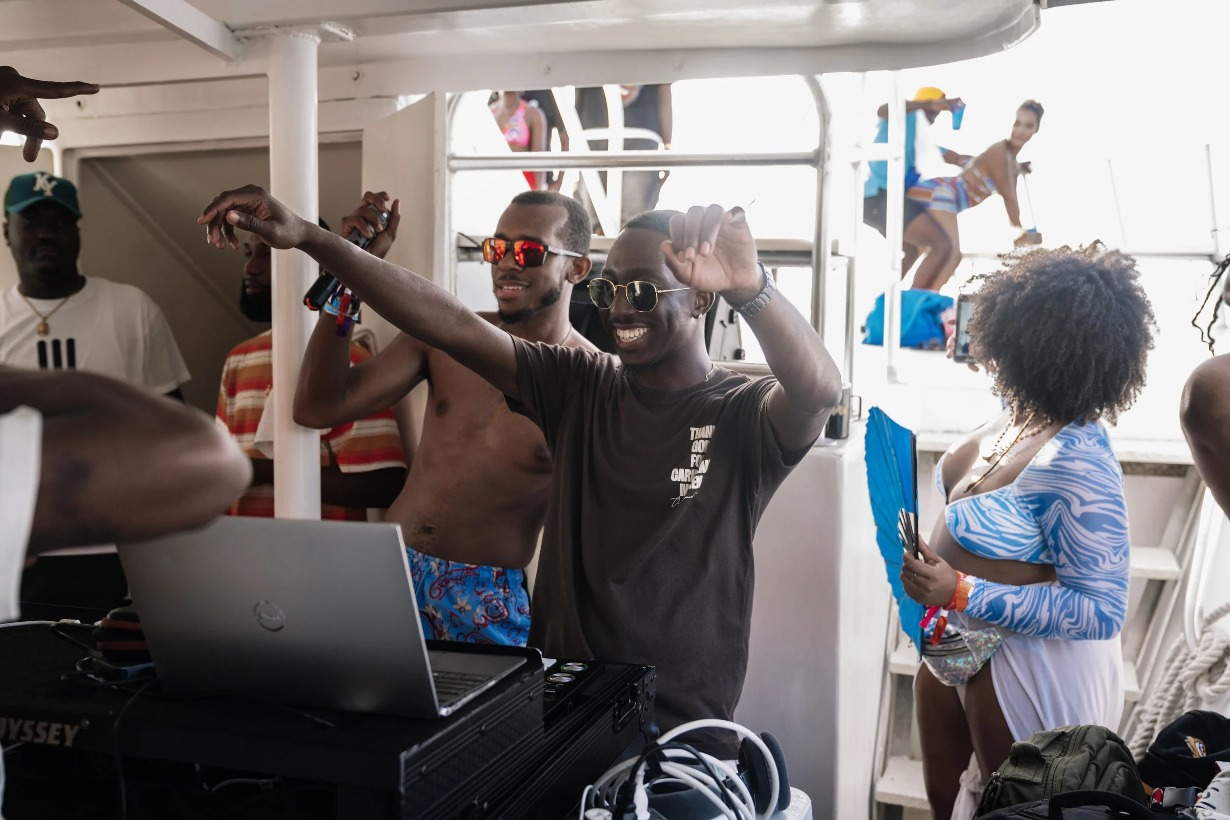 Group of people enjoying a party on a boat, with a man DJing and others dancing and socializing, some dressed in swimwear.