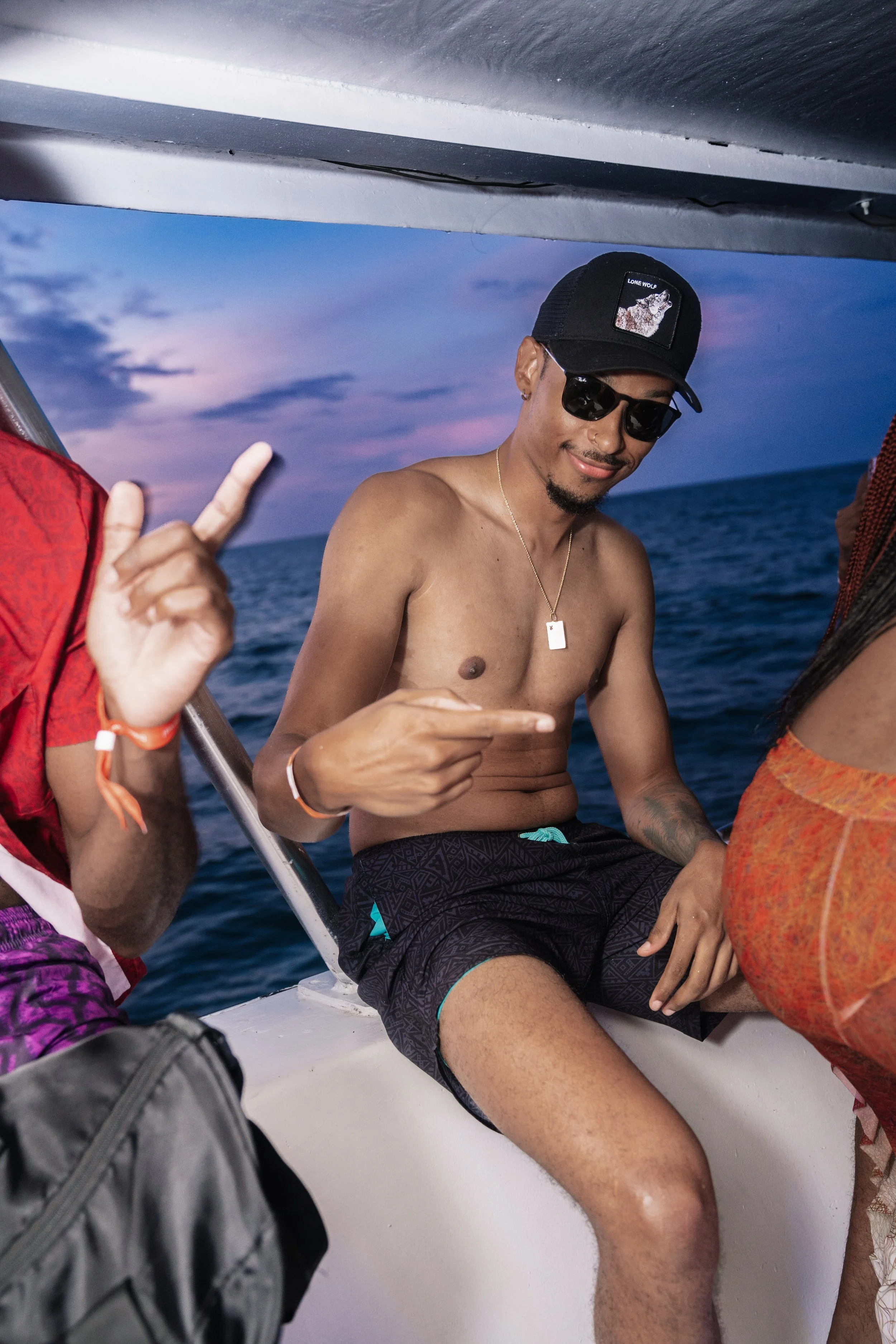 A shirtless man wearing sunglasses, a black cap, and shorts, sitting on a boat with an ocean and sunset sky in the background, smiling and pointing at himself.