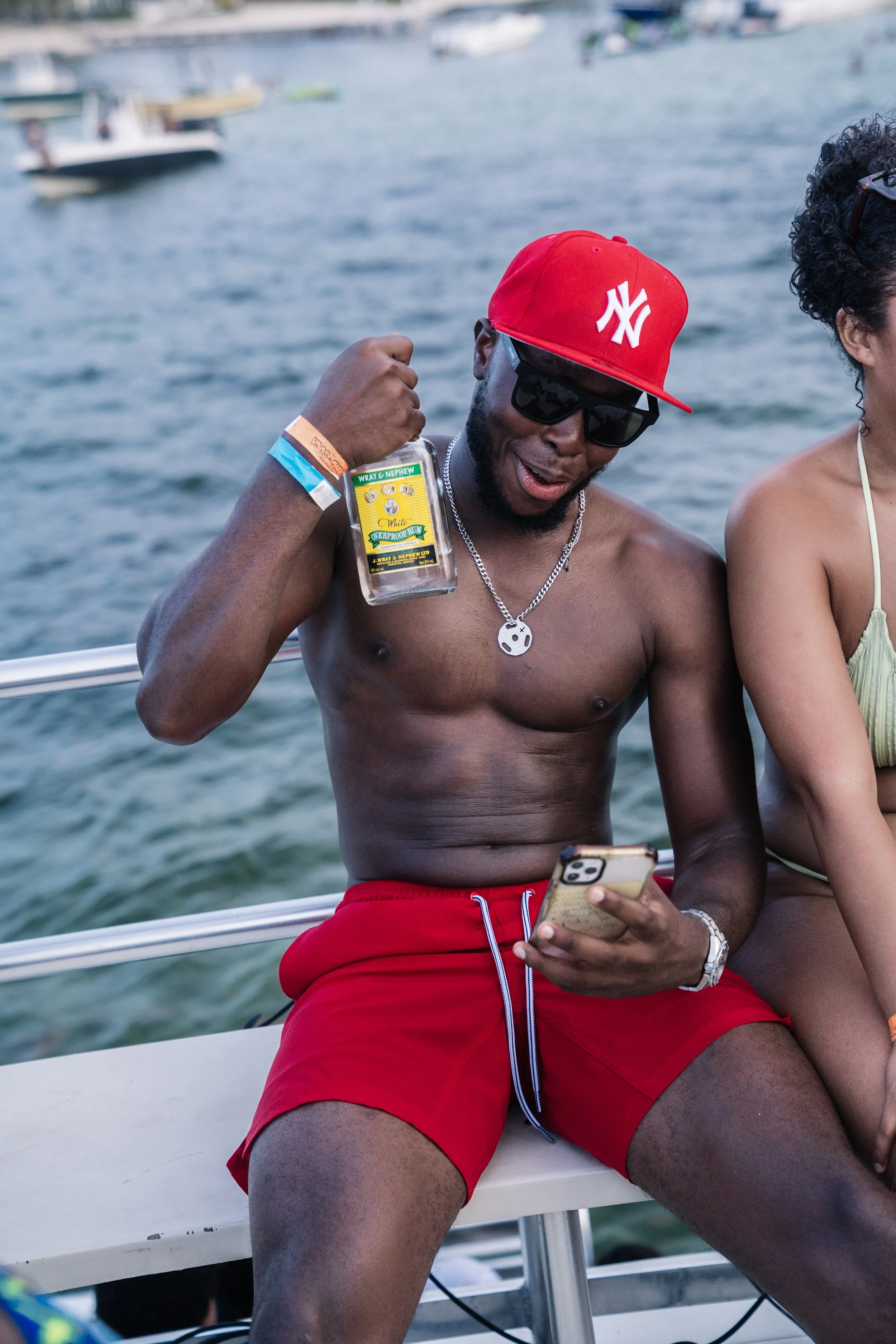 A shirtless man wearing a red cap, sunglasses, and red shorts, sitting on a boat beside a woman, holding a phone and a bottle of alcohol, with boats and water in the background.