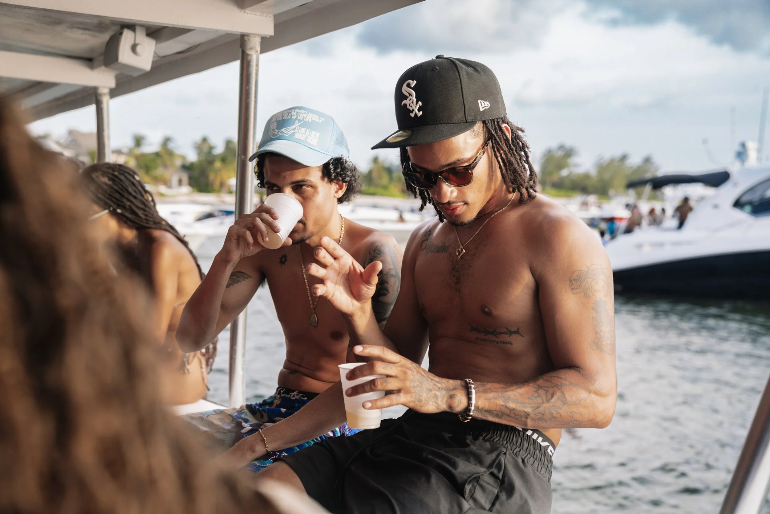 Two shirtless men with tattoos, one wearing a black baseball cap and sunglasses, and the other with a blue cap, sitting on a boat near a marina, drinking from disposable cups.