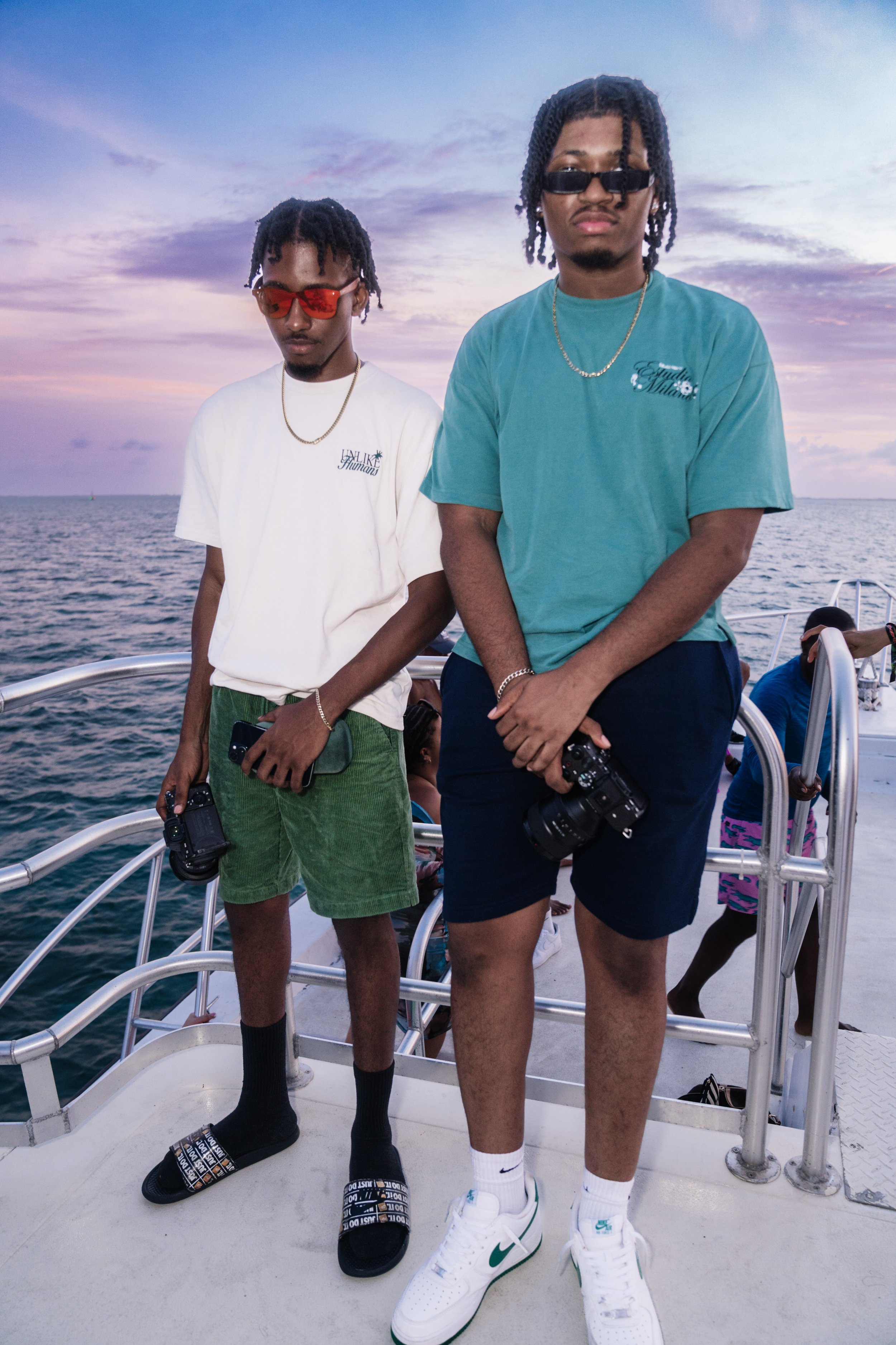 Two young men standing on a boat tijdens sunset. One wears a white t-shirt, green shorts, black slide sandals, red sunglasses, and jewelry, holding a camera and phone. The other is dressed in a teal t-shirt, black shorts, white sneakers, glasses, and