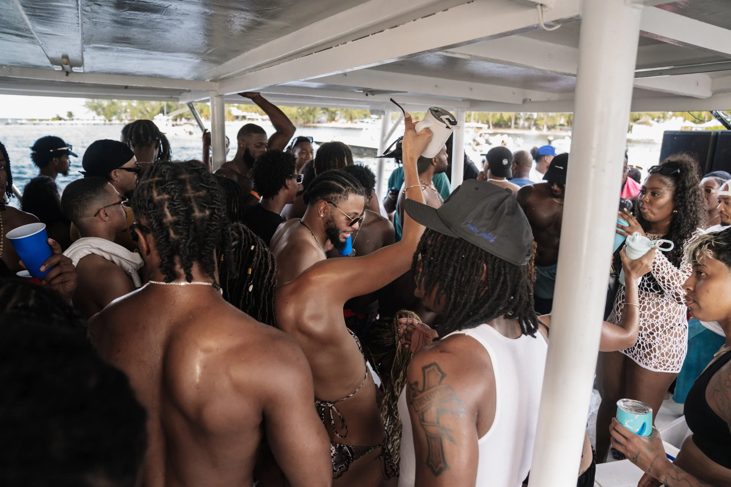 A group of people dancing and socializing on a boat during daytime, with water and trees visible in the background.