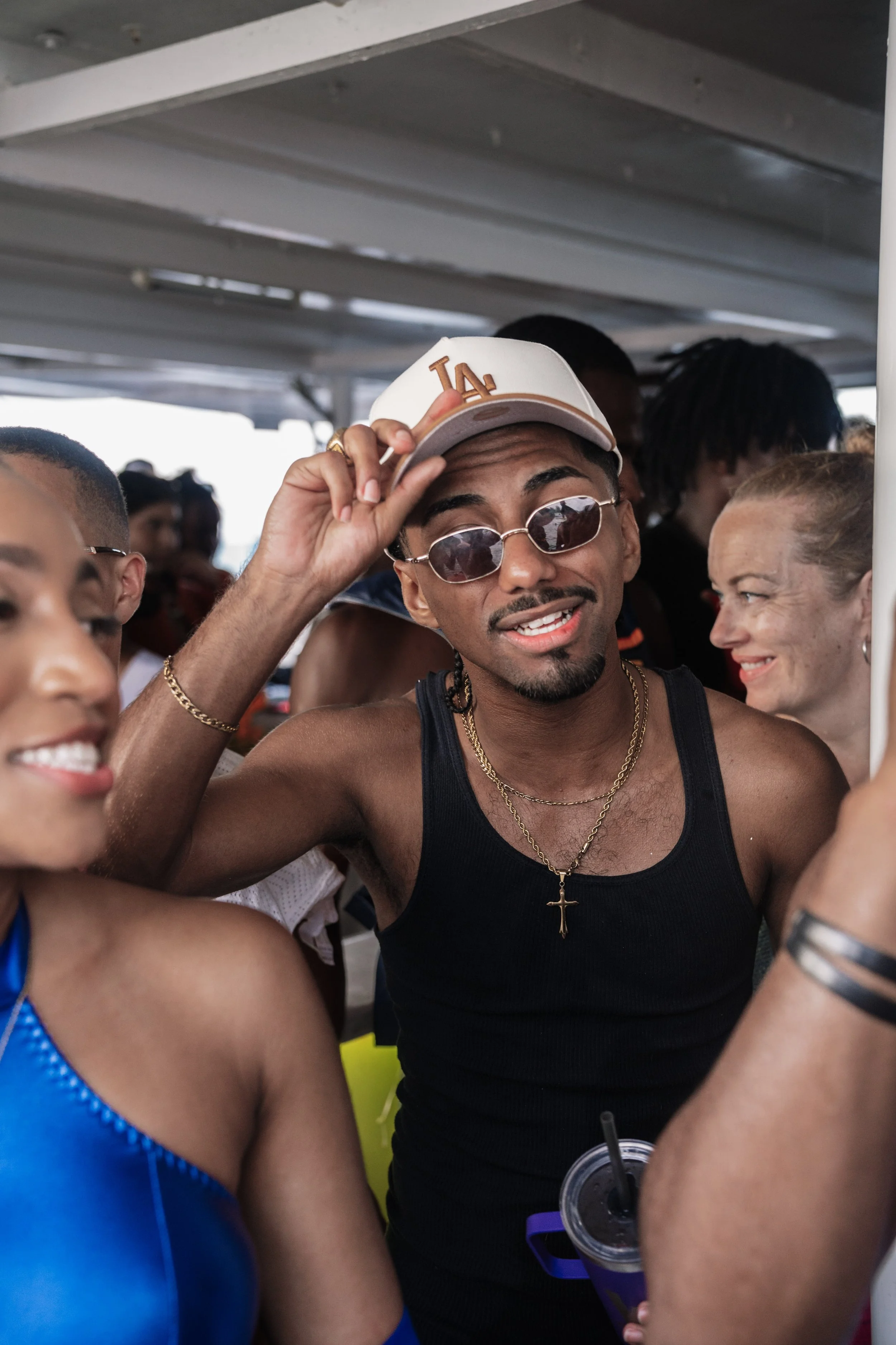A group of people enjoying themselves on a boat or ferry. The central person is a man wearing a LA cap, sunglasses, a black tank top, and gold jewelry. He is smiling and adjusting his cap. Other people around him are smiling and talking.