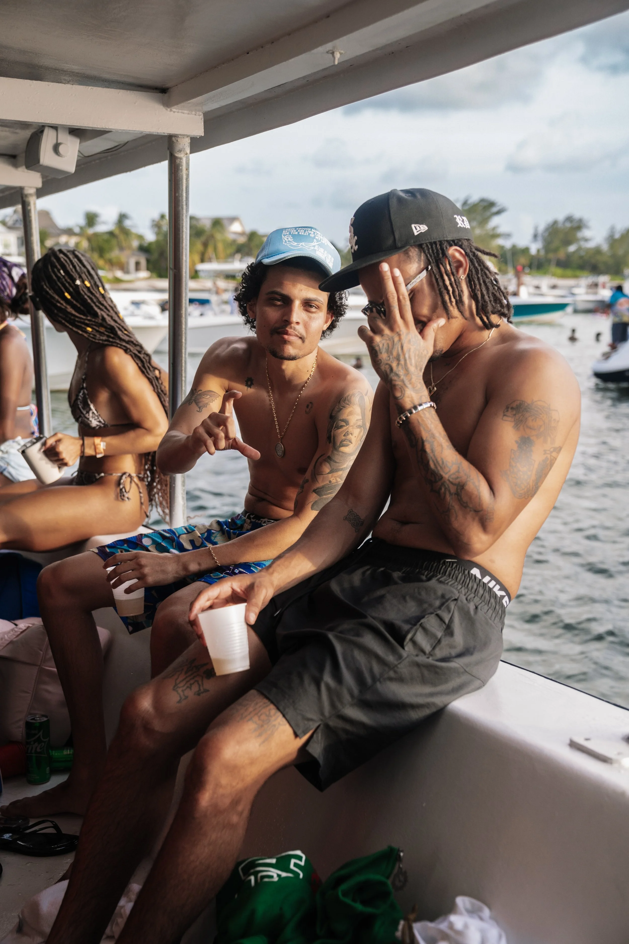 Two shirtless men with tattoos and wearing caps sitting on a boat, holding cups, with a waterway and boats in the background.