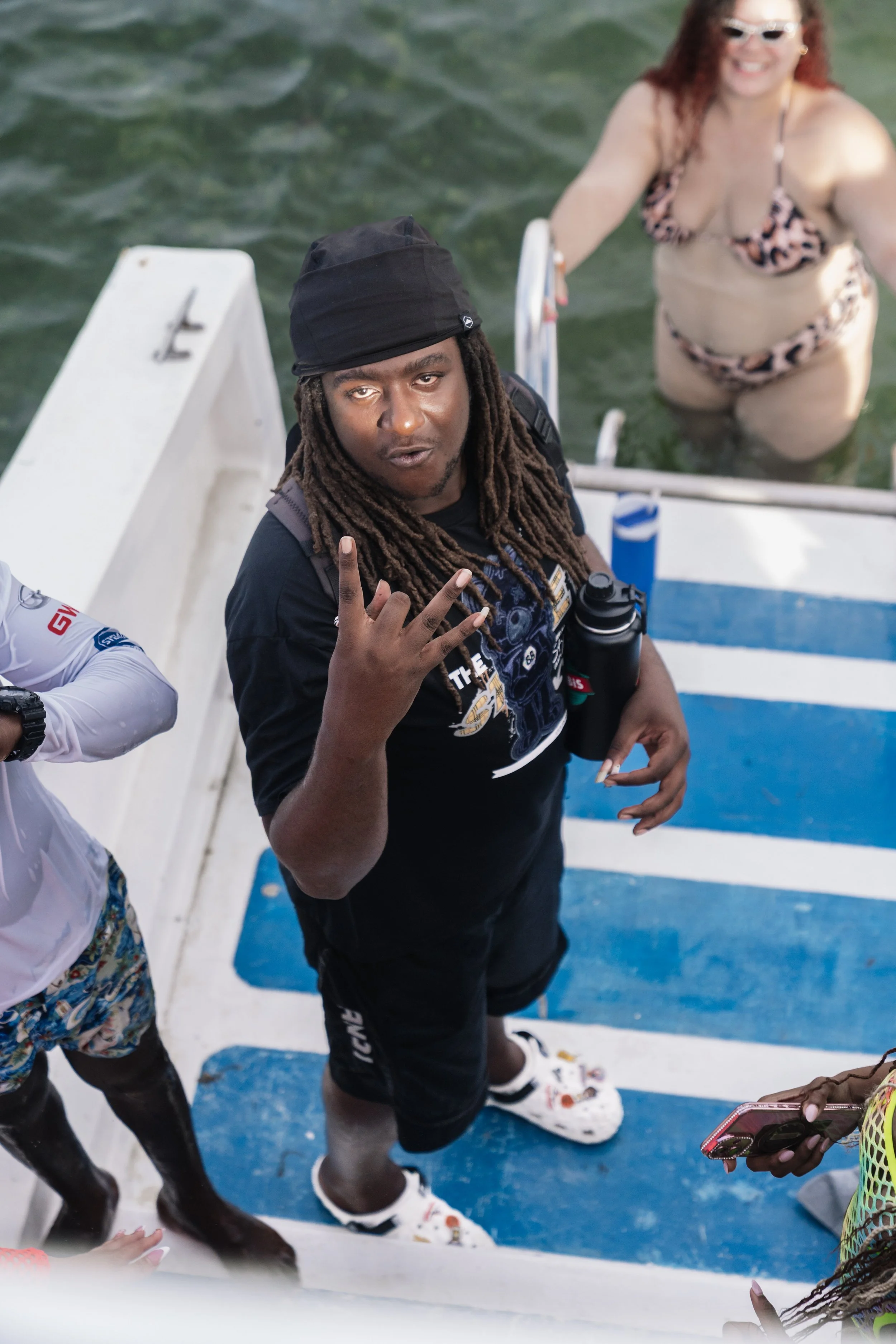 Man with long dreadlocks making a hand gesture while standing on a boat, with a woman in a bikini in the water behind him.
